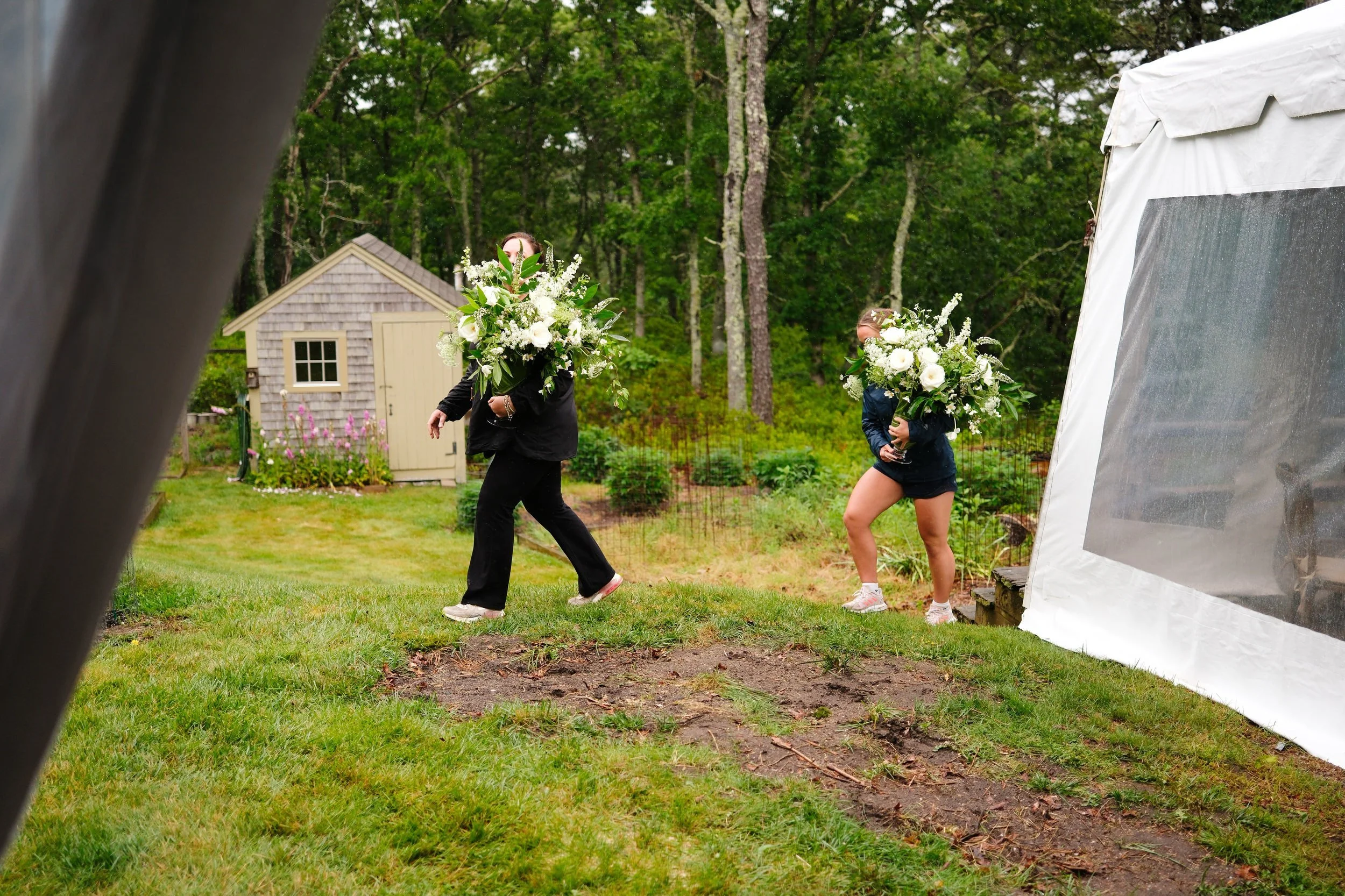Two women carrying large bouquets of white flowers walk on a grassy backyard with a small shed, trees, and a garden in the background, partially visible from an outdoor tent.