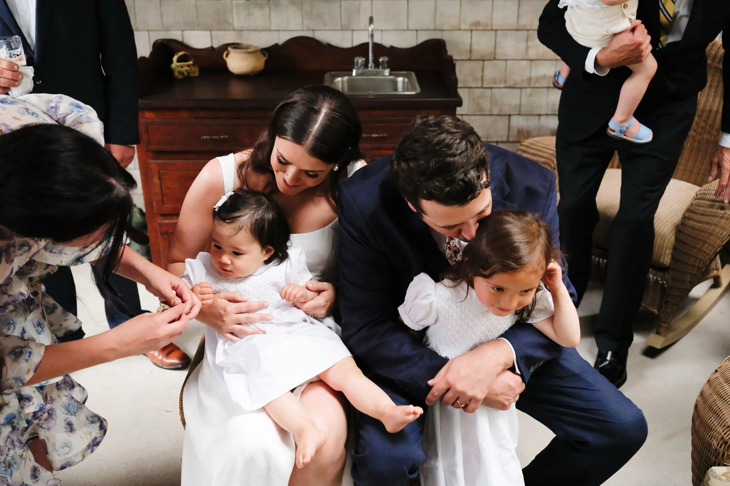 A woman and a young girl in white dresses are seated, with the woman holding a smaller child in a white dress. Two men in suits are crouched down next to them, one holding a girl in a white dress. Multiple people are gathered around, some partially v