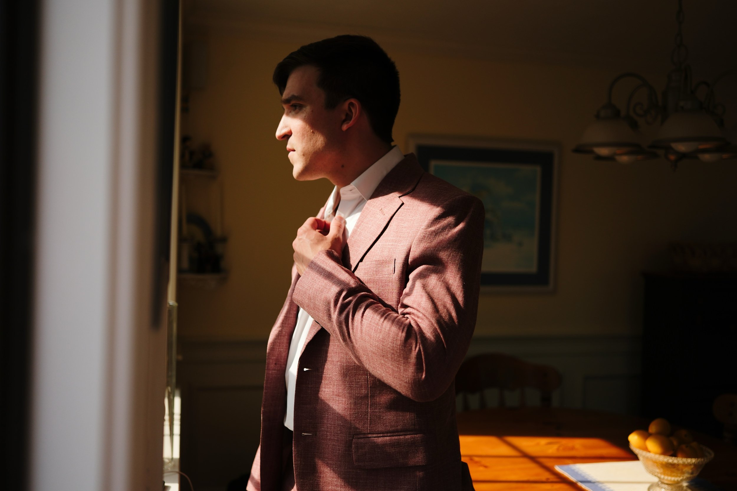 A man in a pinkish-brown suit adjusting his collar while looking out a window, with warm lighting and a blurred background of a dining room.