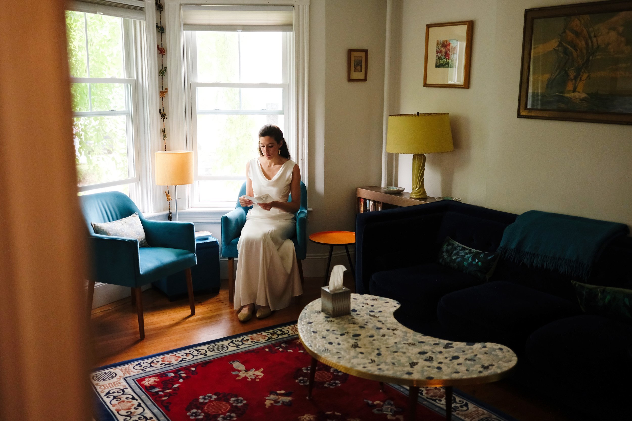 A woman in a white dress sitting in a teal armchair near a window, reading a piece of paper in a cozy living room with artwork on the walls, a yellow lamp on a side table, and a marble coffee table with tissues.