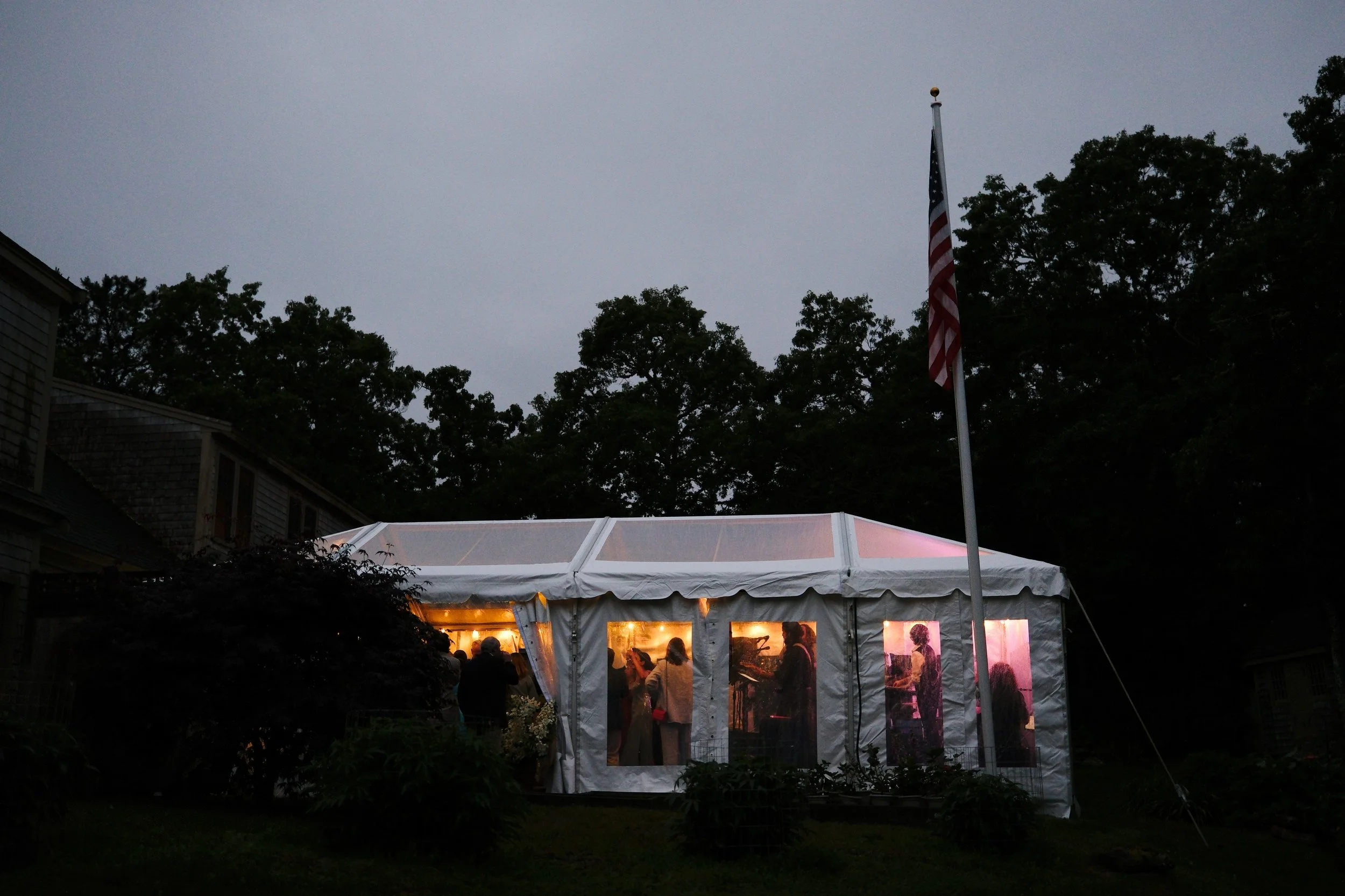 A white event tent illuminated from within, set up outdoors at dusk, with several people inside and a flagpole with the American flag nearby.