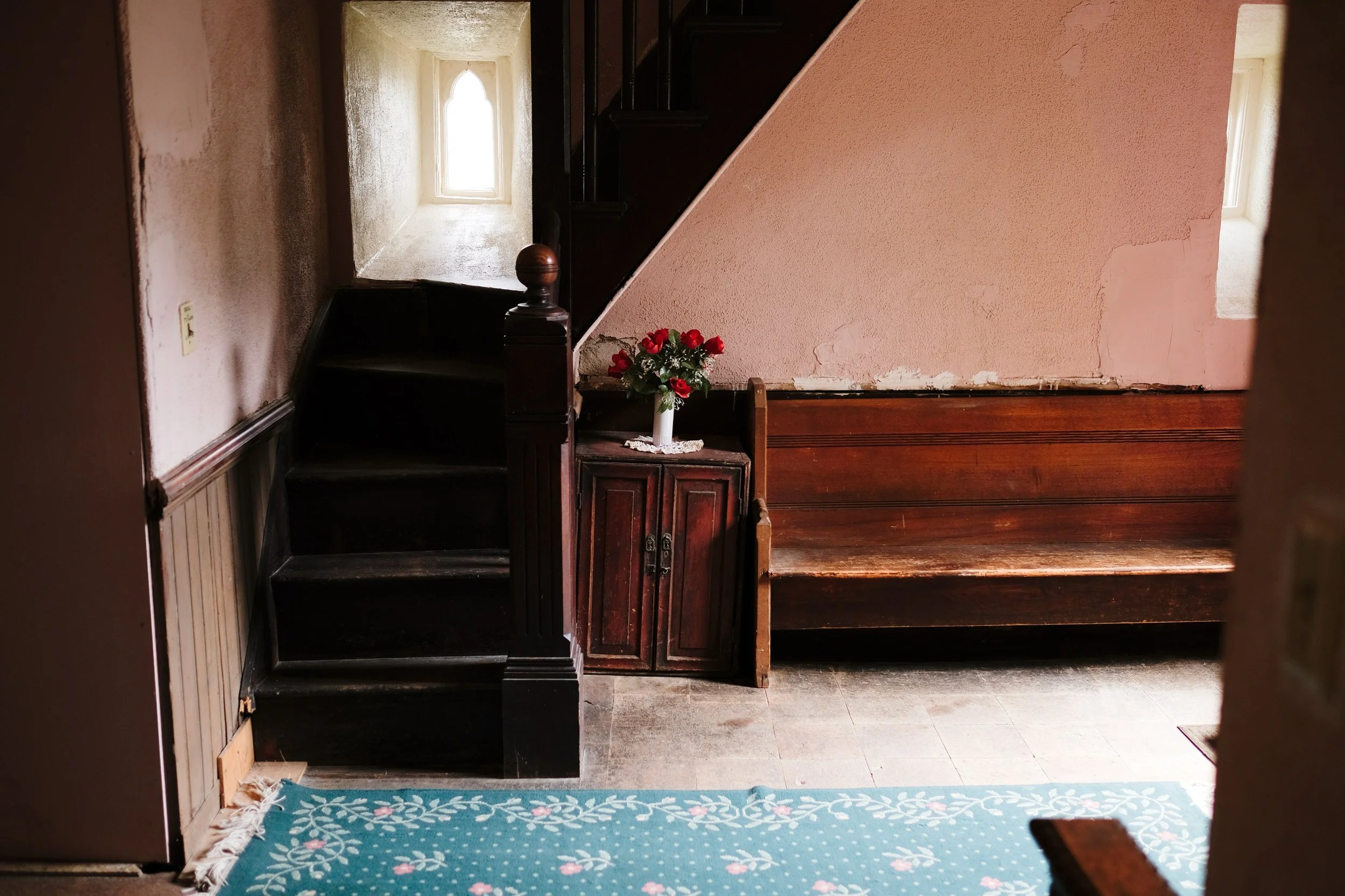 Interior view of a small room with a staircase, a bench, a flower vase with red roses, and two small windows letting in natural light.