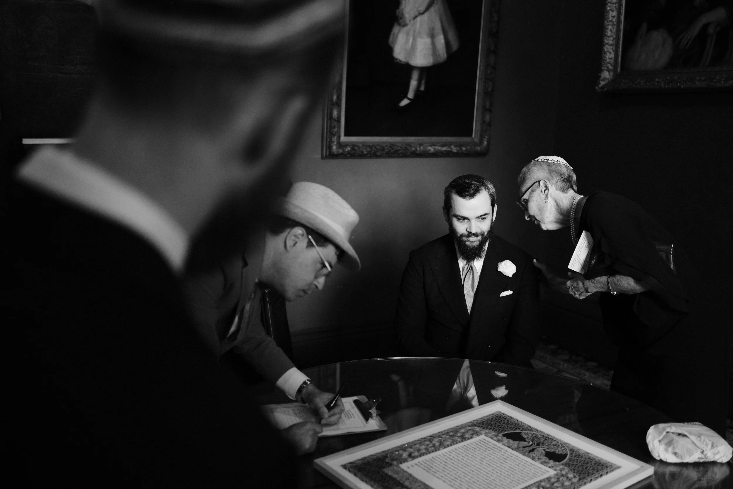 Black and white photo of four people at a meeting or discussion around a table in a room with framed artwork on the wall. One person is taking notes, and the others are engaged in conversation.