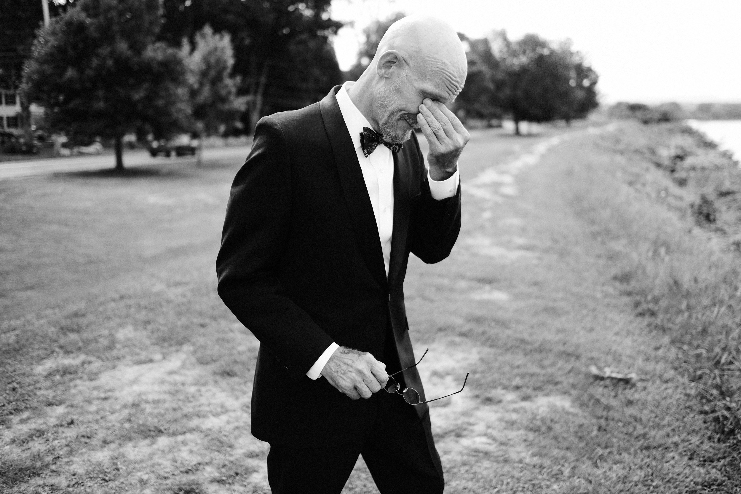 An elderly man in a tuxedo with a bow tie, holding glasses, standing outdoors near a river, with a distressed expression, covering his face with one hand.