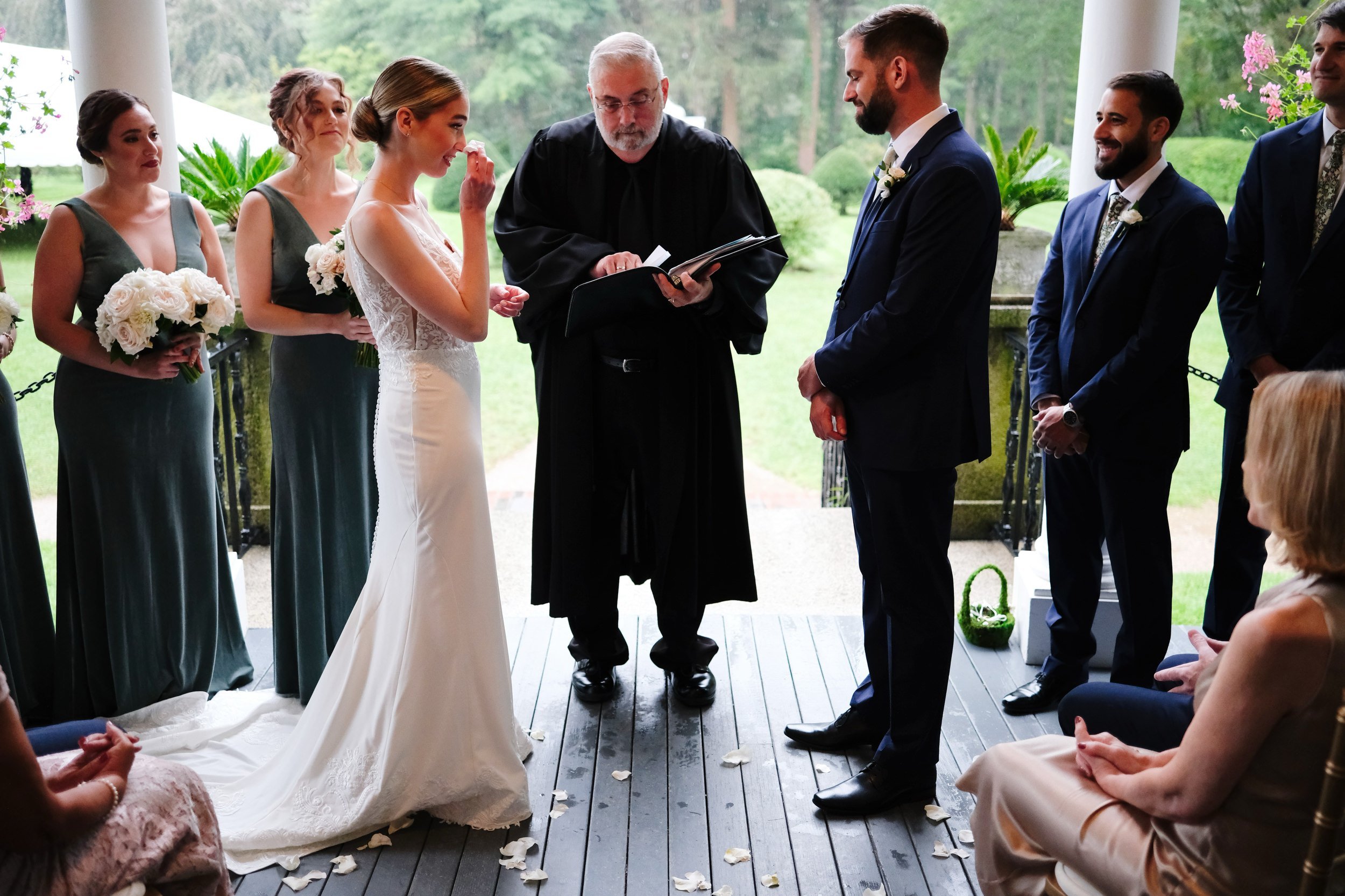 A wedding ceremony taking place outdoors under a covered porch, with a bride and groom facing each other, surrounded by bridesmaids and groomsmen. The bride is dressed in a white gown holding a bouquet, and the groom in a dark suit. An officiant in b