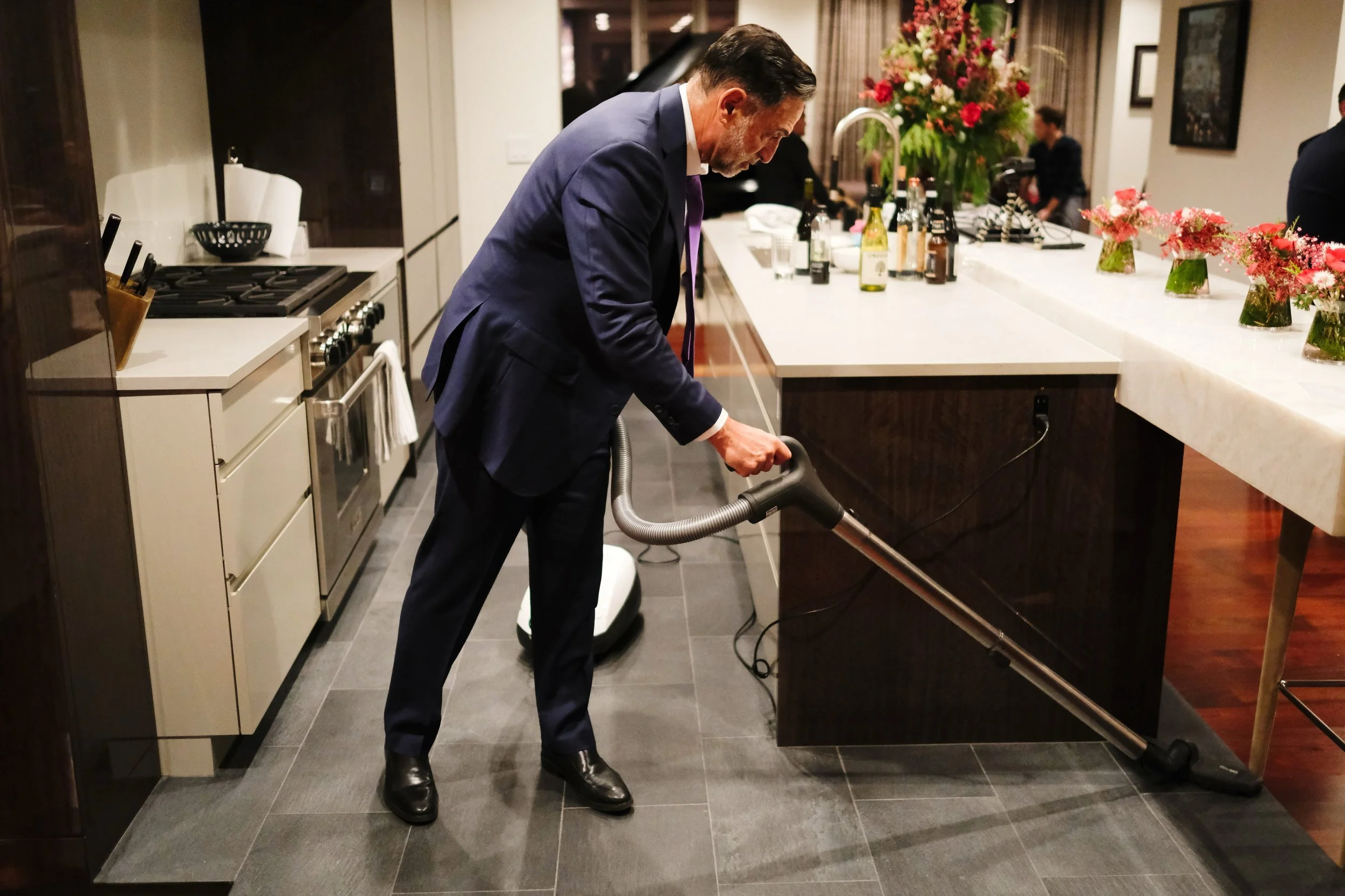 A man in a dark suit using a vacuum cleaner to clean the kitchen floor in a modern kitchen with white cabinetry, a large granite island, and flower arrangements.