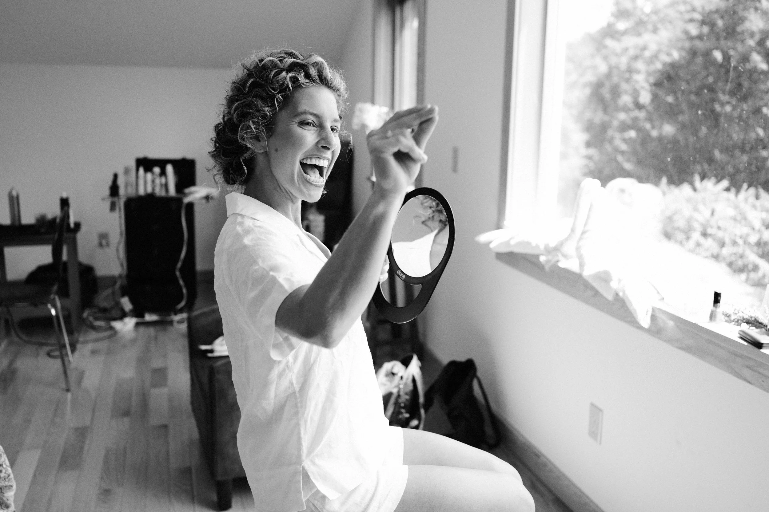 A woman with curly hair and wearing a white shirt is sitting near a window, smiling and holding a mirror in one hand. The room appears to be a casual living space with furniture and objects in the background.