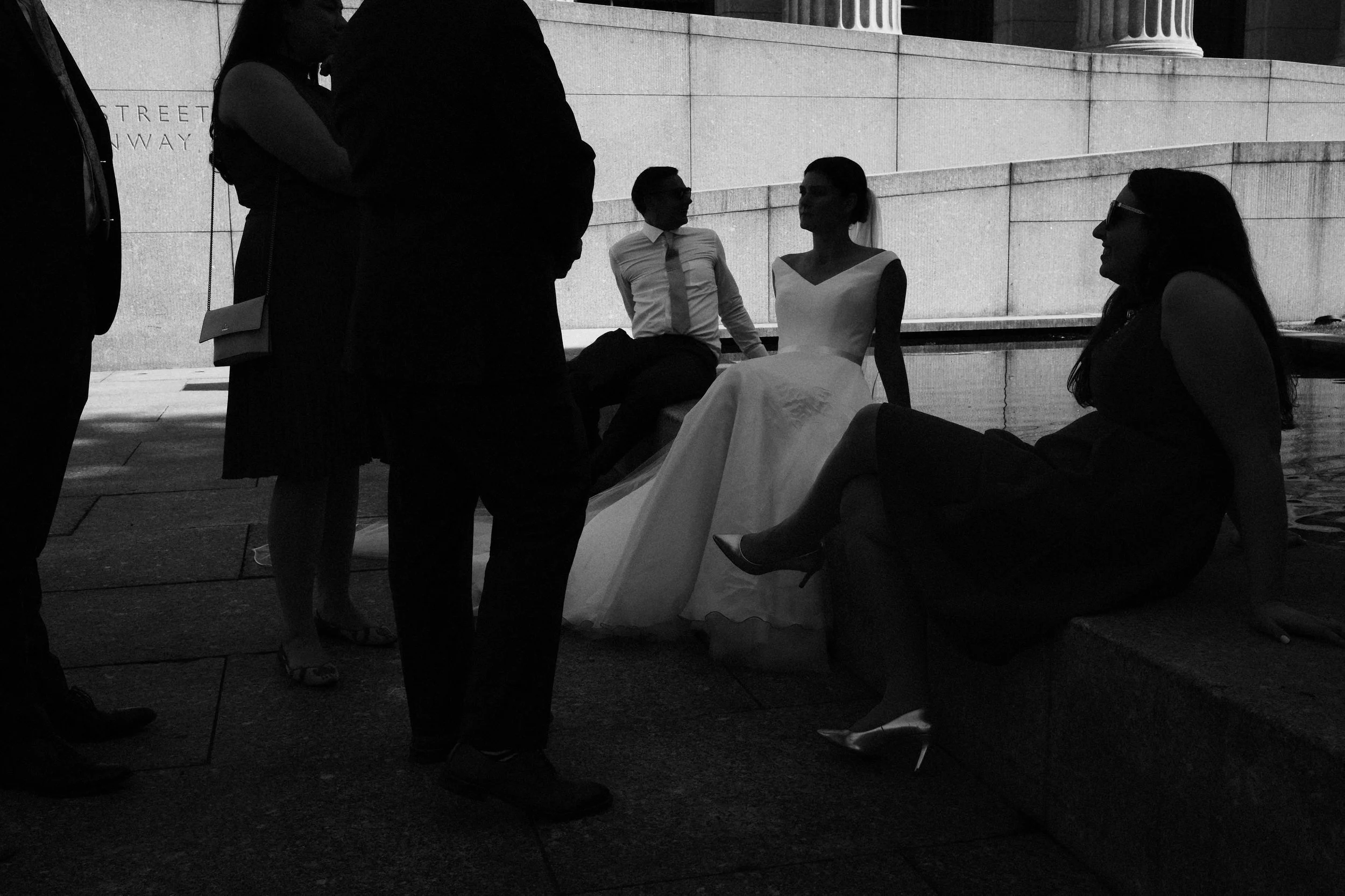 Group of people in formal attire, black and white photo, some seated and some standing, in an outdoor urban setting near a building with stone steps.