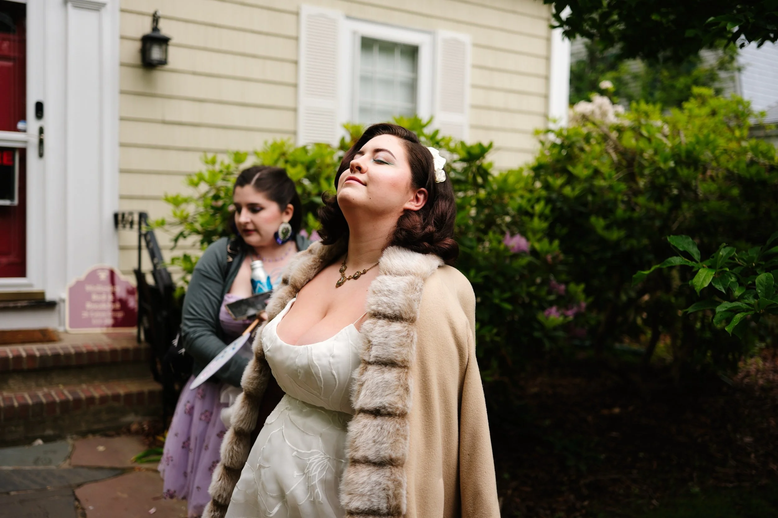 A woman with dark hair, wearing a cream-colored dress and a fur-lined coat, stands with her eyes closed and a serene expression. Behind her, another woman with dark hair in a bun, wearing a lavender dress with floral patterns and a dark jacket, appea