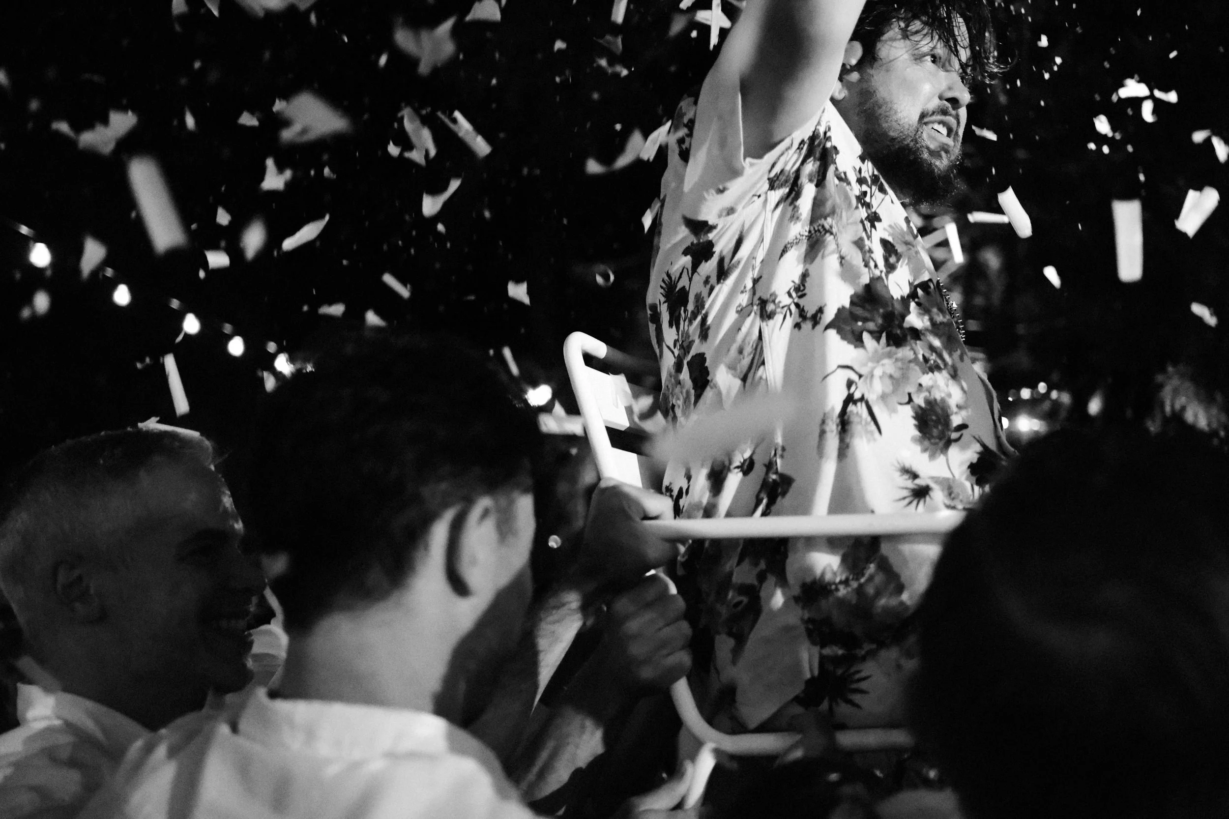 A man with a beard and curly hair, wearing a floral shirt, is sitting on a chair surrounded by people. The scene is dark with confetti falling around them, suggesting a celebration.