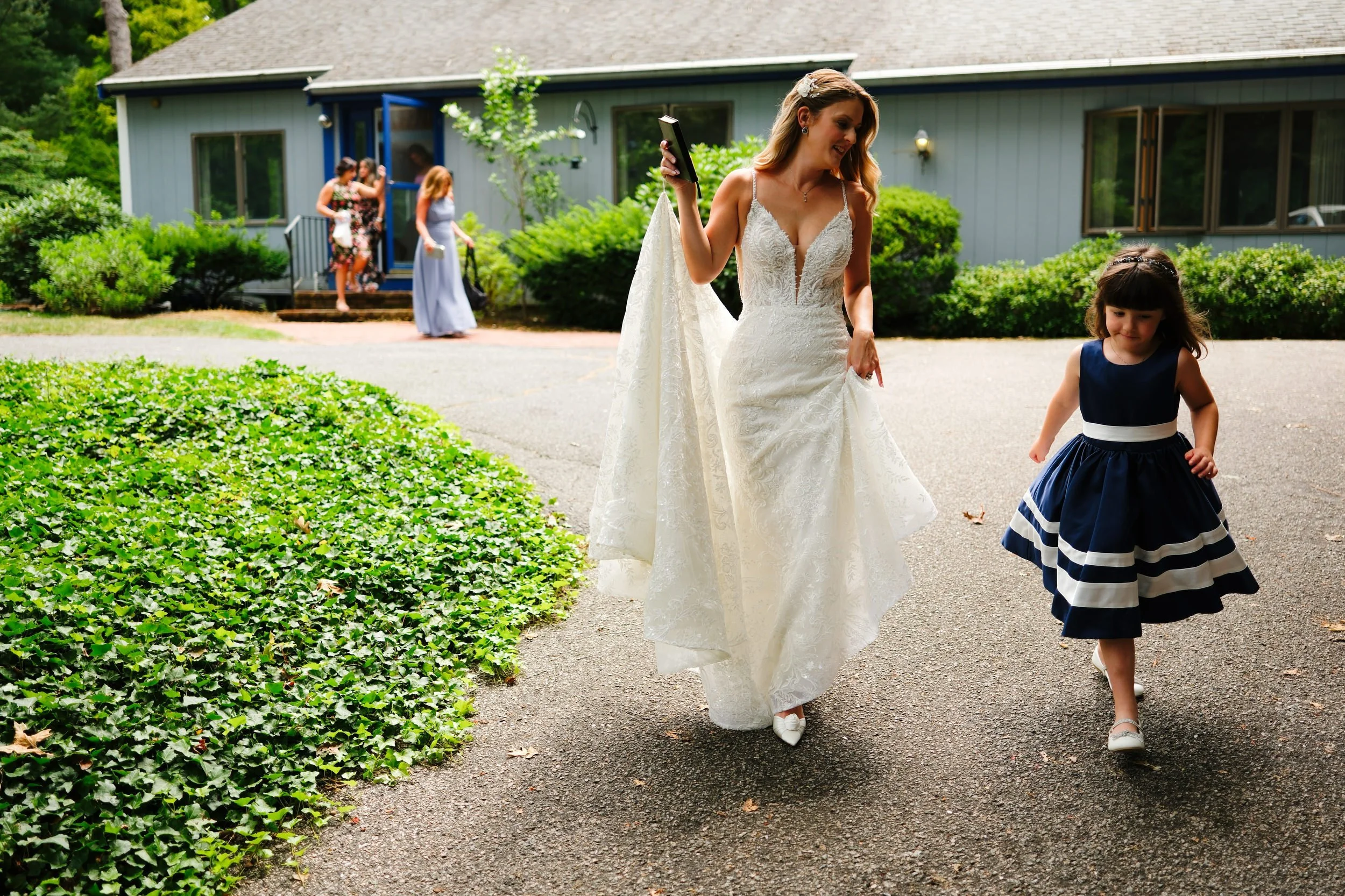 A bride in a white wedding dress walking outdoors with a young girl in a navy blue dress. Other women are seen in the background near a light blue house with lush greenery.