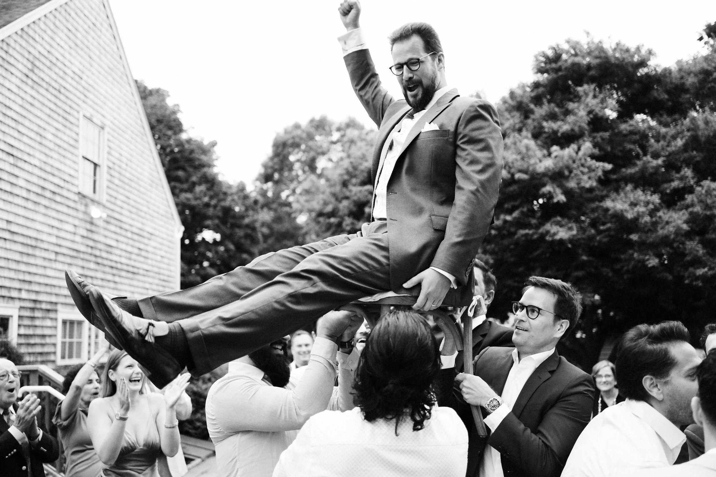 A man in a suit is being lifted into the air on a chair during a celebration, surrounded by smiling and clapping people outdoors, with trees and a house in the background.