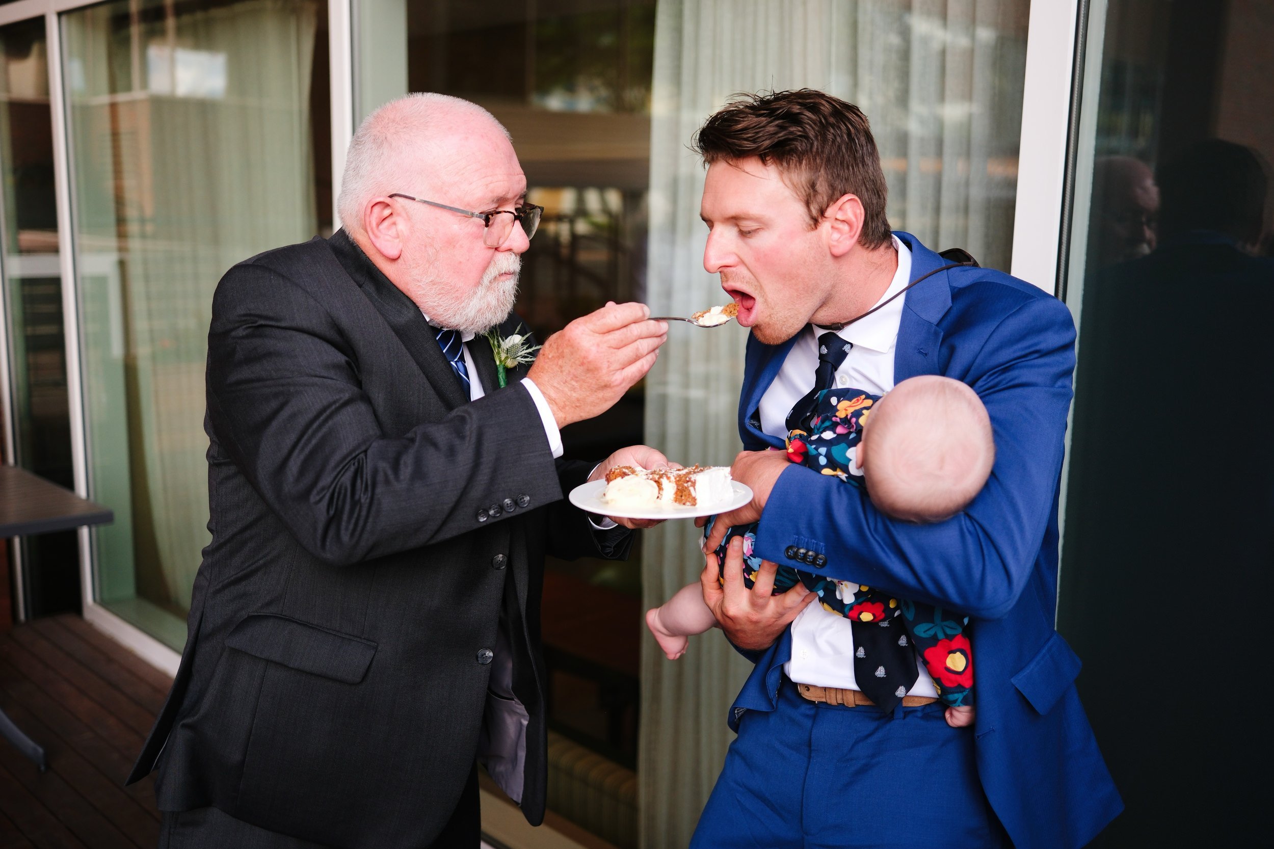 Man in blue suit holding baby and feeding wedding cake to an older man in black suit