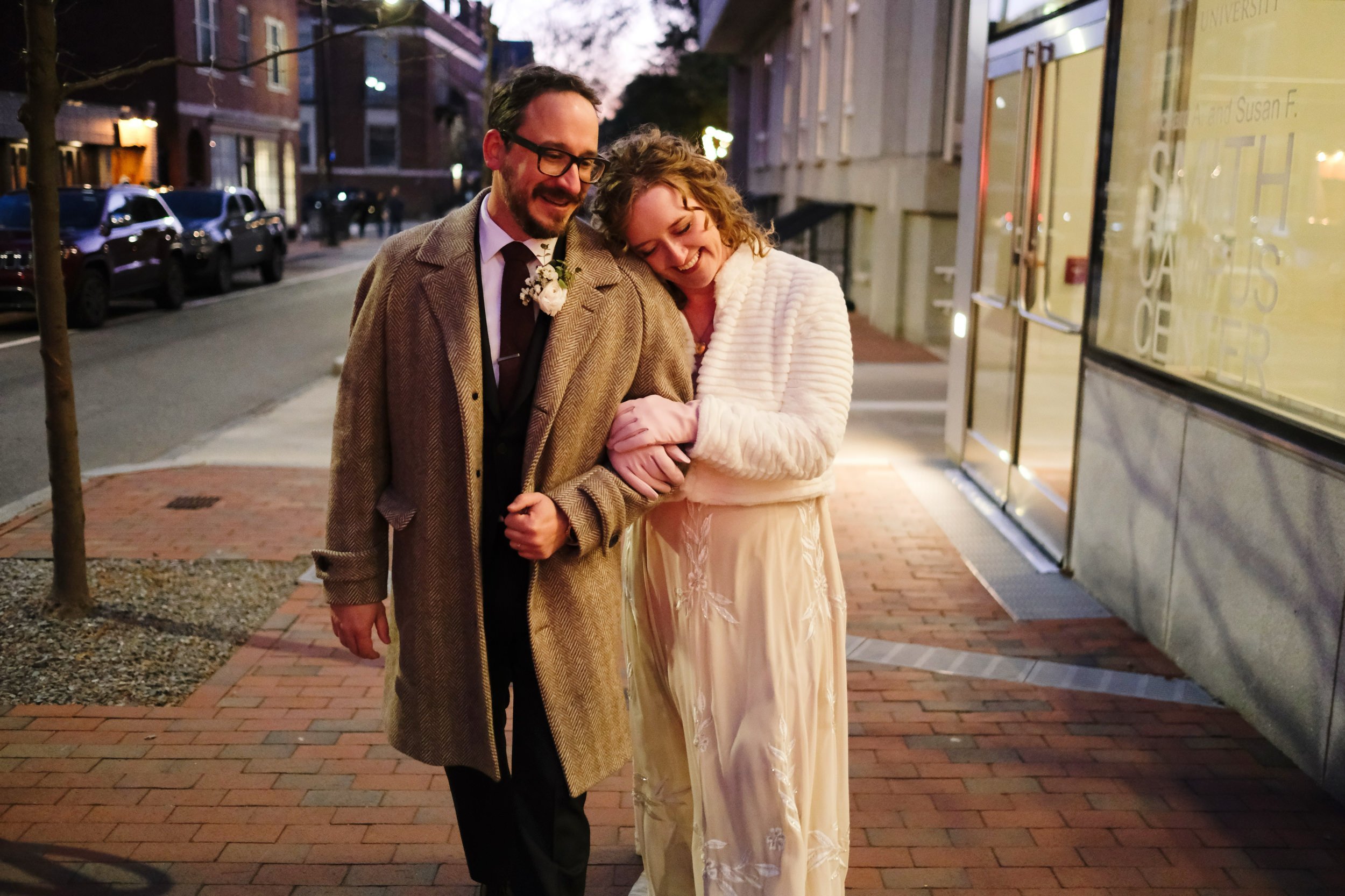 A happy couple walking arm in arm on a city sidewalk during dusk, smiling and enjoying each other's company.