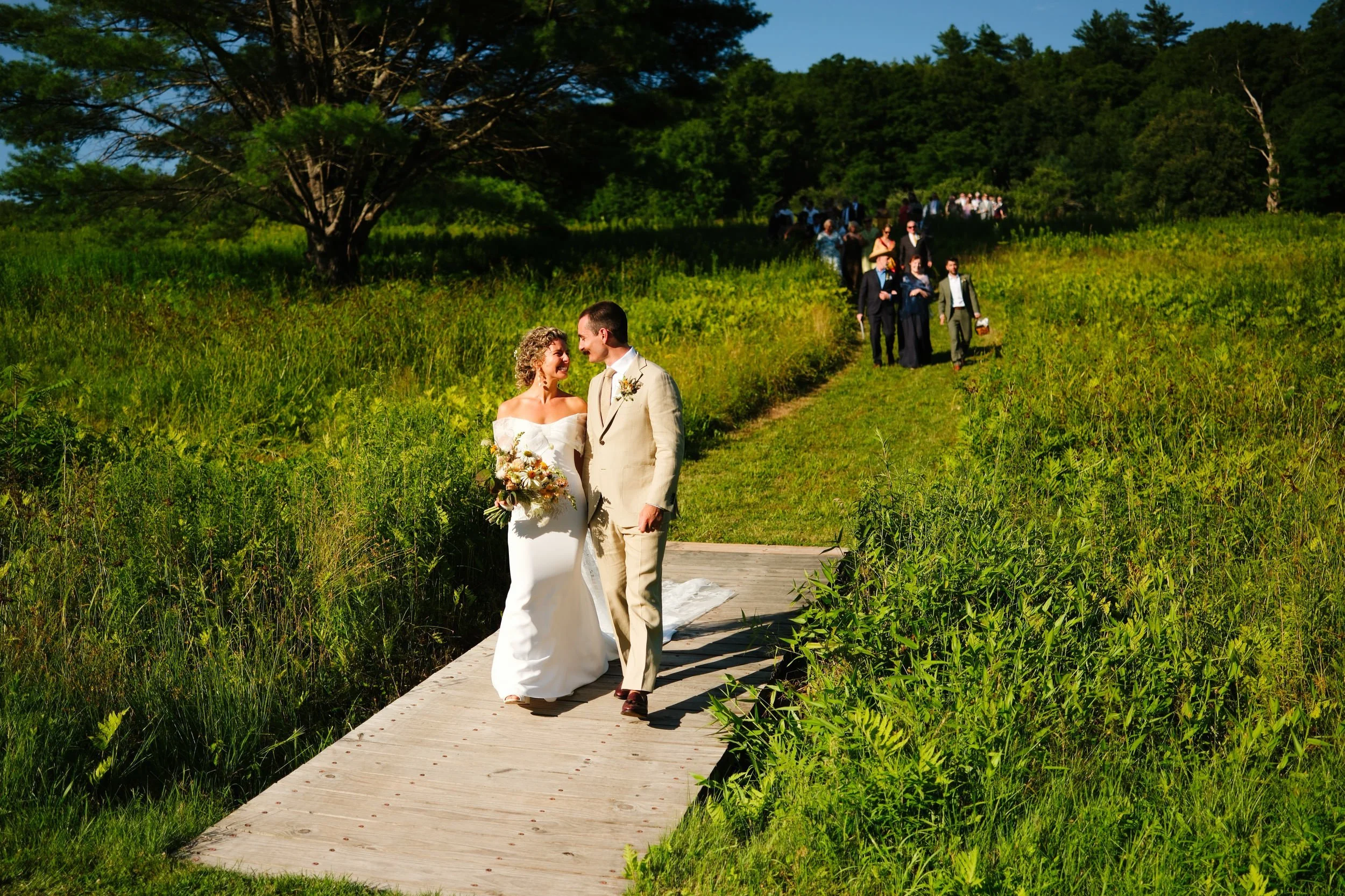 Bride and groom walking on a wooden path in a lush green outdoor setting during their wedding, with wedding guests in the background.