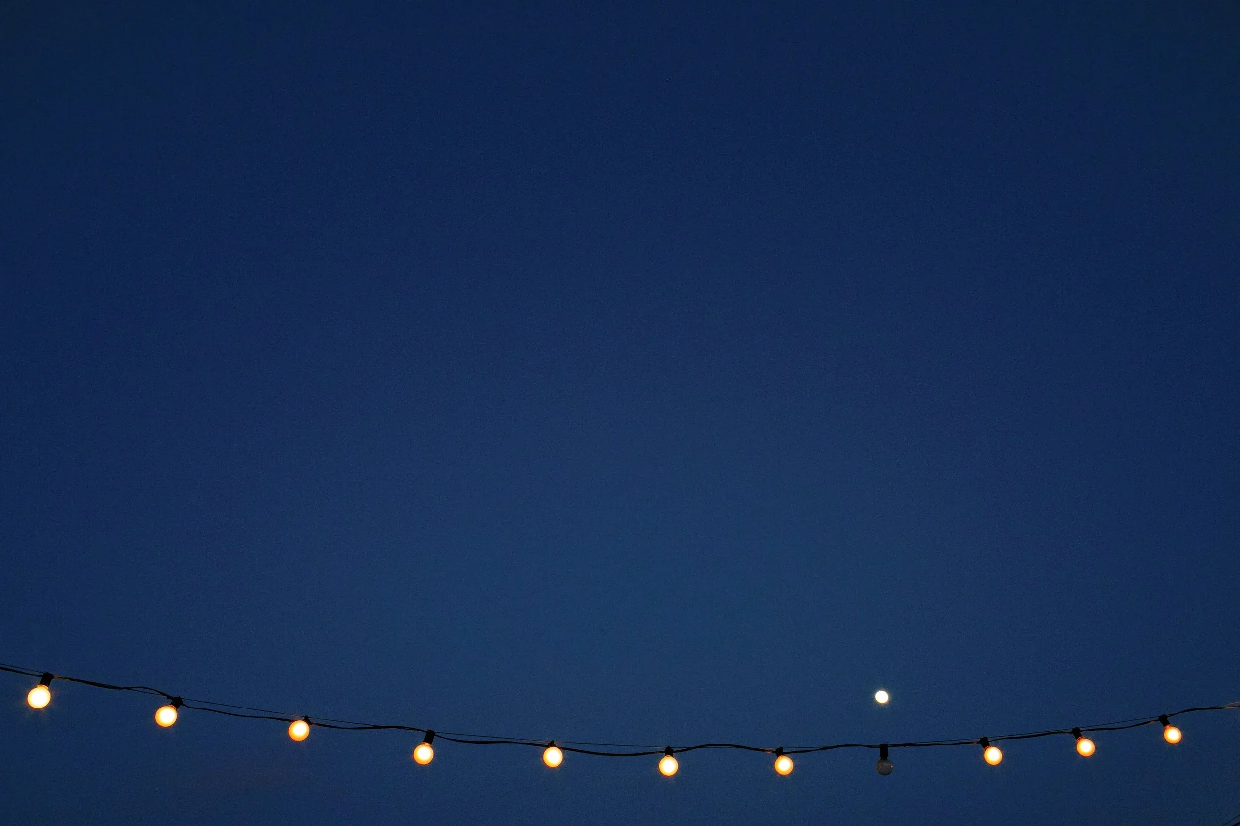 String of hanging outdoor fairy lights against a dark blue evening sky with a visible moon at a wedding reception.