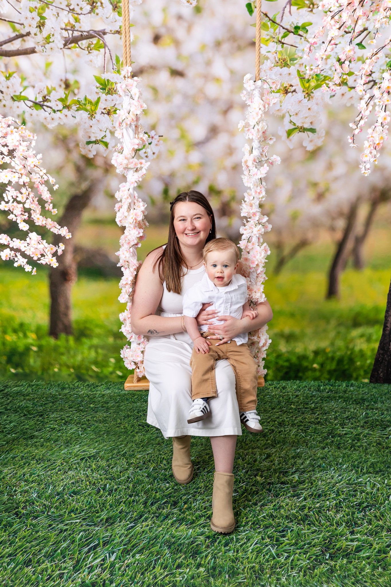 A woman and a young boy sitting on a swing decorated with pink flowers, outdoors with flowering trees in the background.