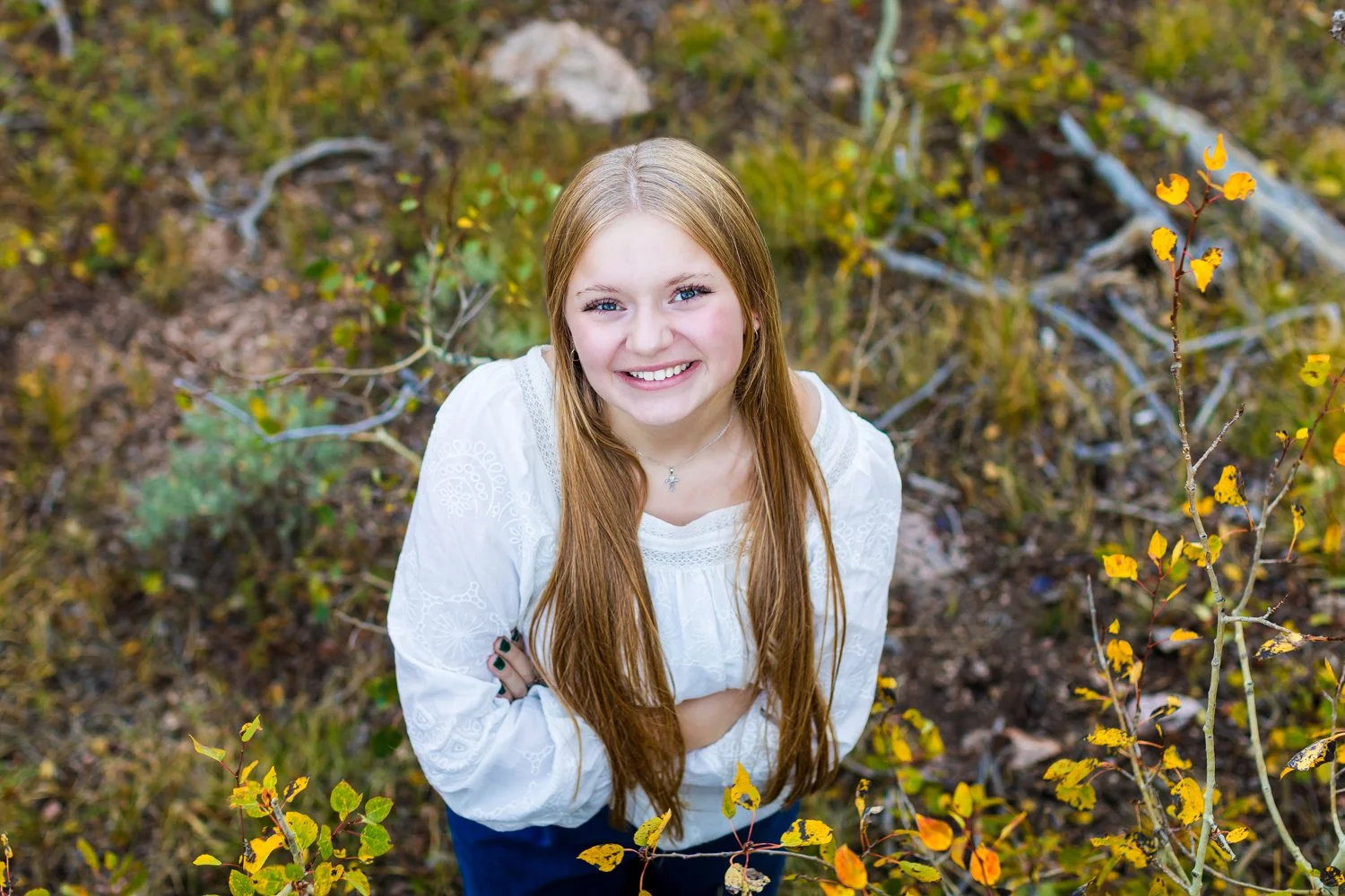 A young woman with long red hair smiling outdoors in a forest or park with yellow and green foliage, looking up at the camera.