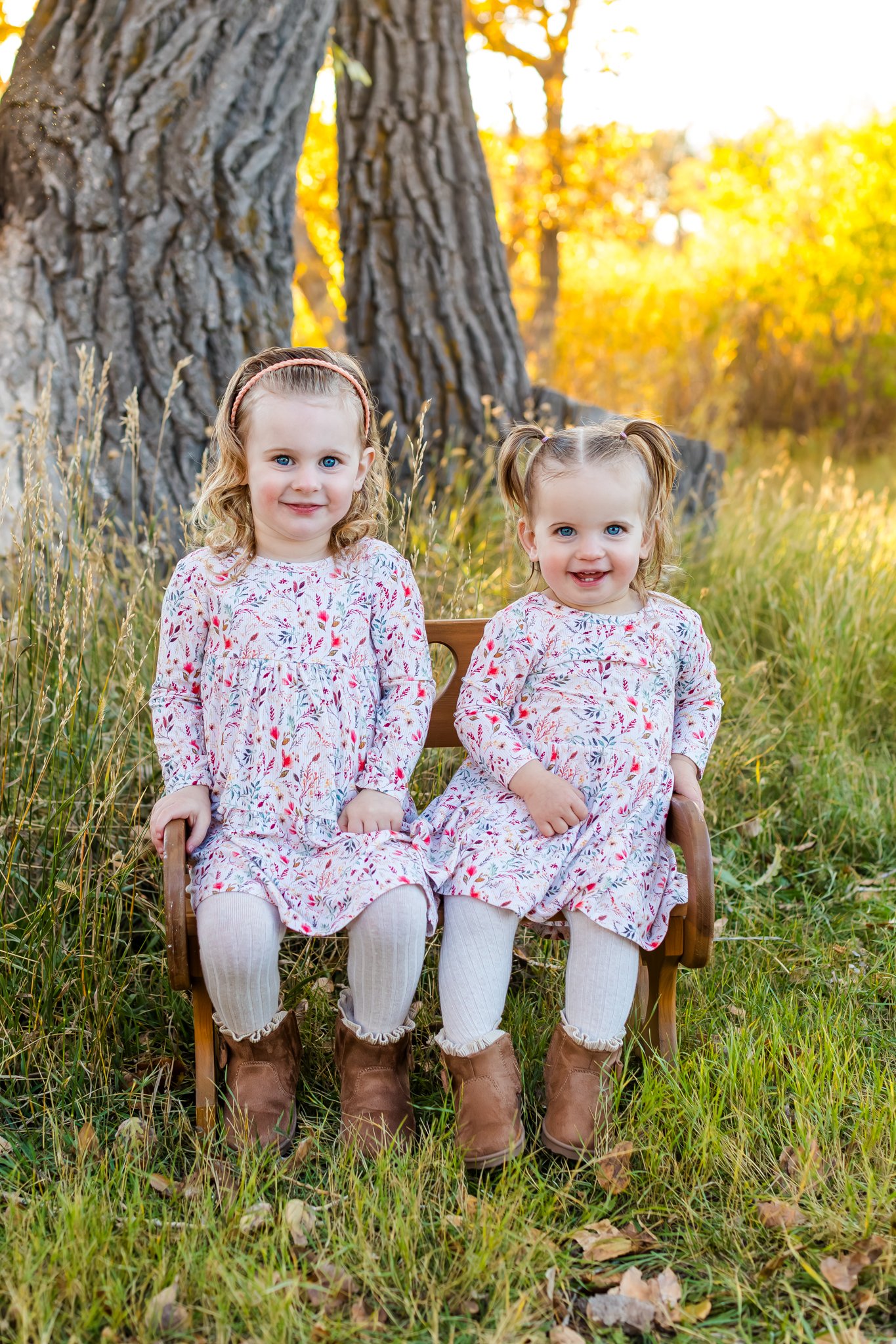 Two young girls with light skin and blonde hair sitting on a small wooden bench outdoors among tall grass and autumn trees. They are wearing matching floral dresses, white tights, and brown boots, with one girl having her hair in pigtails and the oth