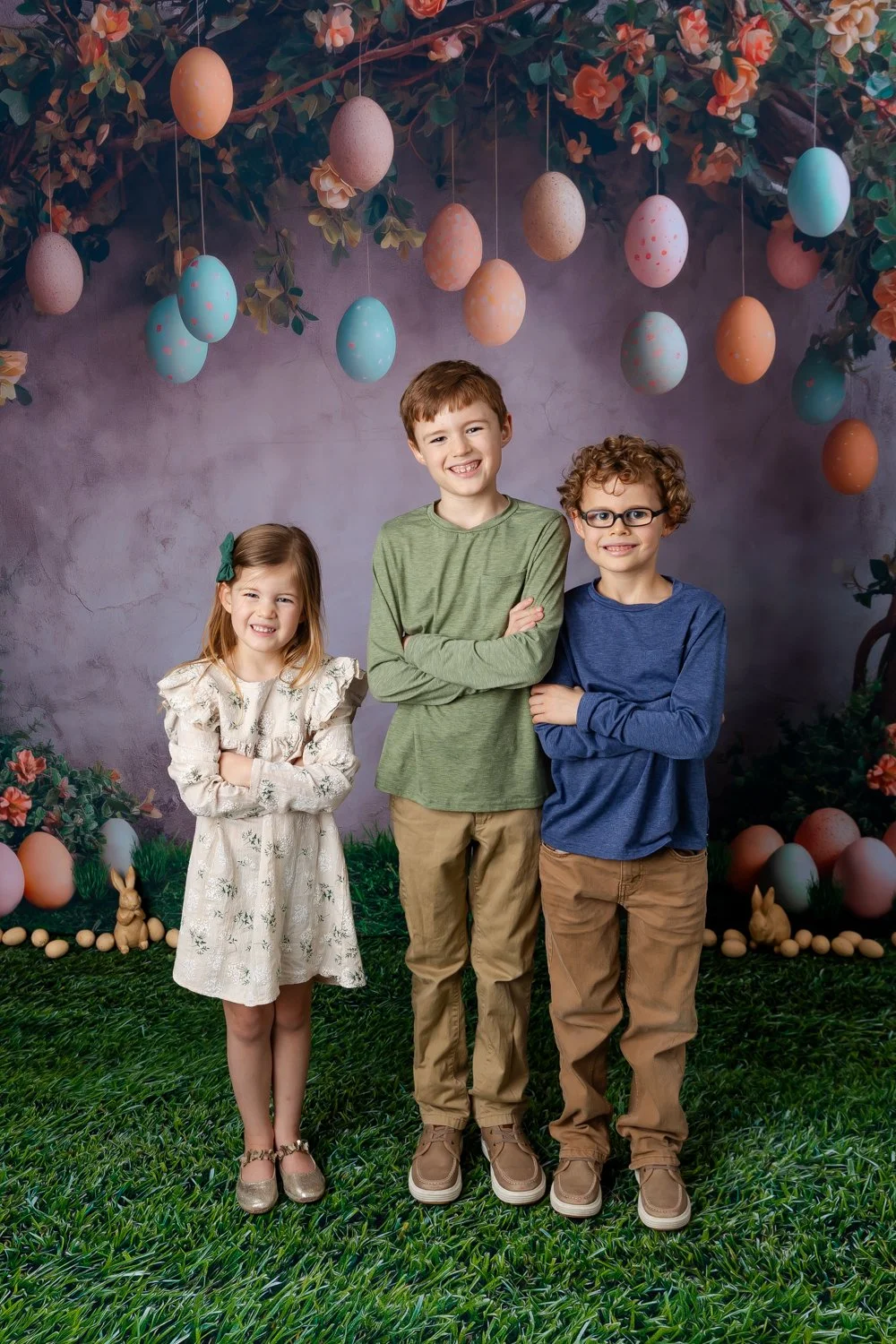 Three children, a girl and two boys, standing on a green grass surface, smiling, with a decorative background featuring hanging pastel-colored eggs, flowers, and Easter decorations.