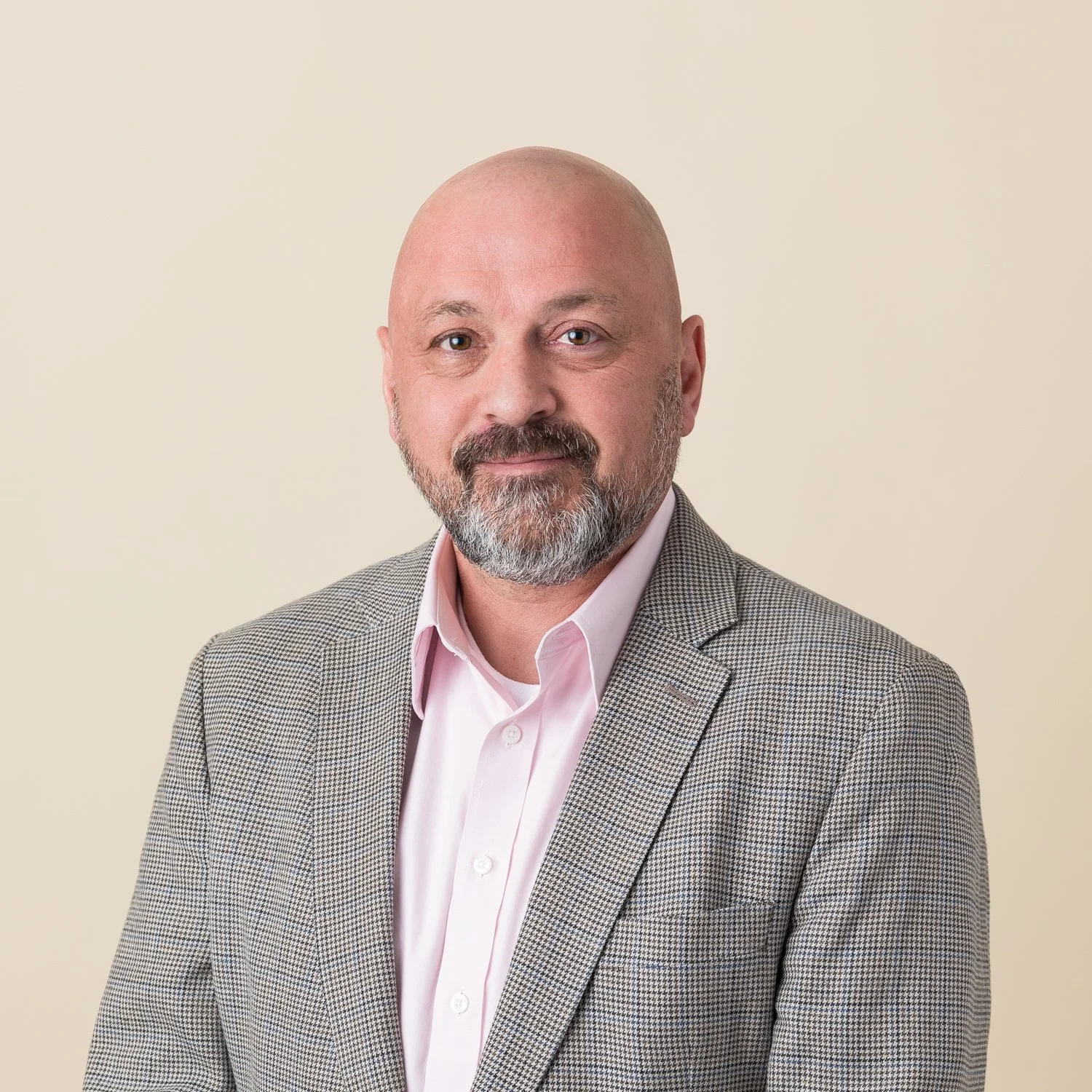 A middle-aged man with a bald head and gray beard wearing a light pink dress shirt and a gray checked blazer, standing against a beige background.