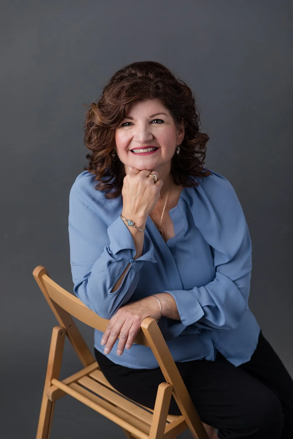 A woman with curly brown hair smiling while sitting on a wooden chair against a gray background, wearing a blue blouse and black pants.
