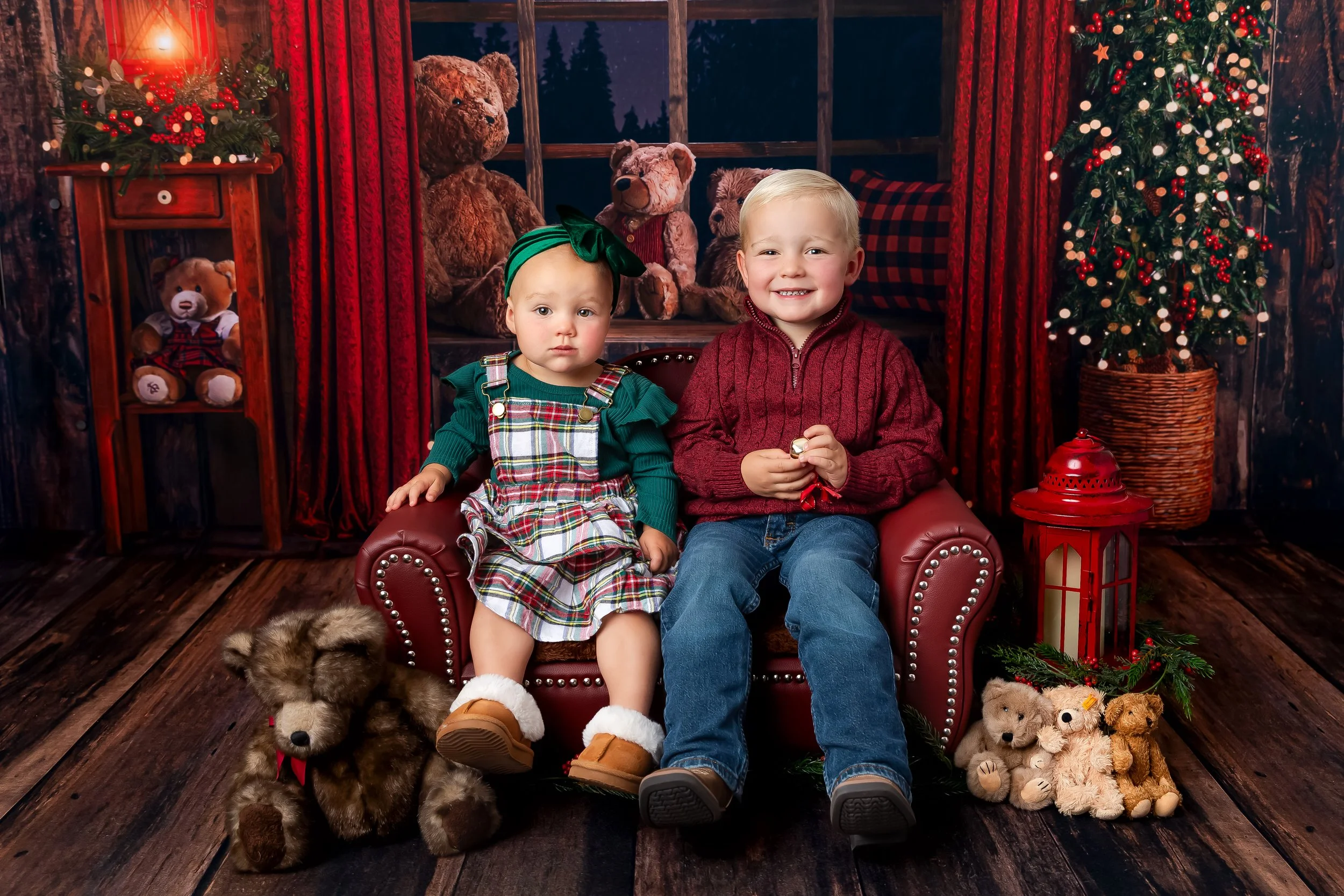 In Cheyenne studio Christmas photos of two young children sitting on a red leather armchair in a Christmas-themed setting. They are surrounded by Christmas decorations including a decorated Christmas tree and stuffed teddy bears.