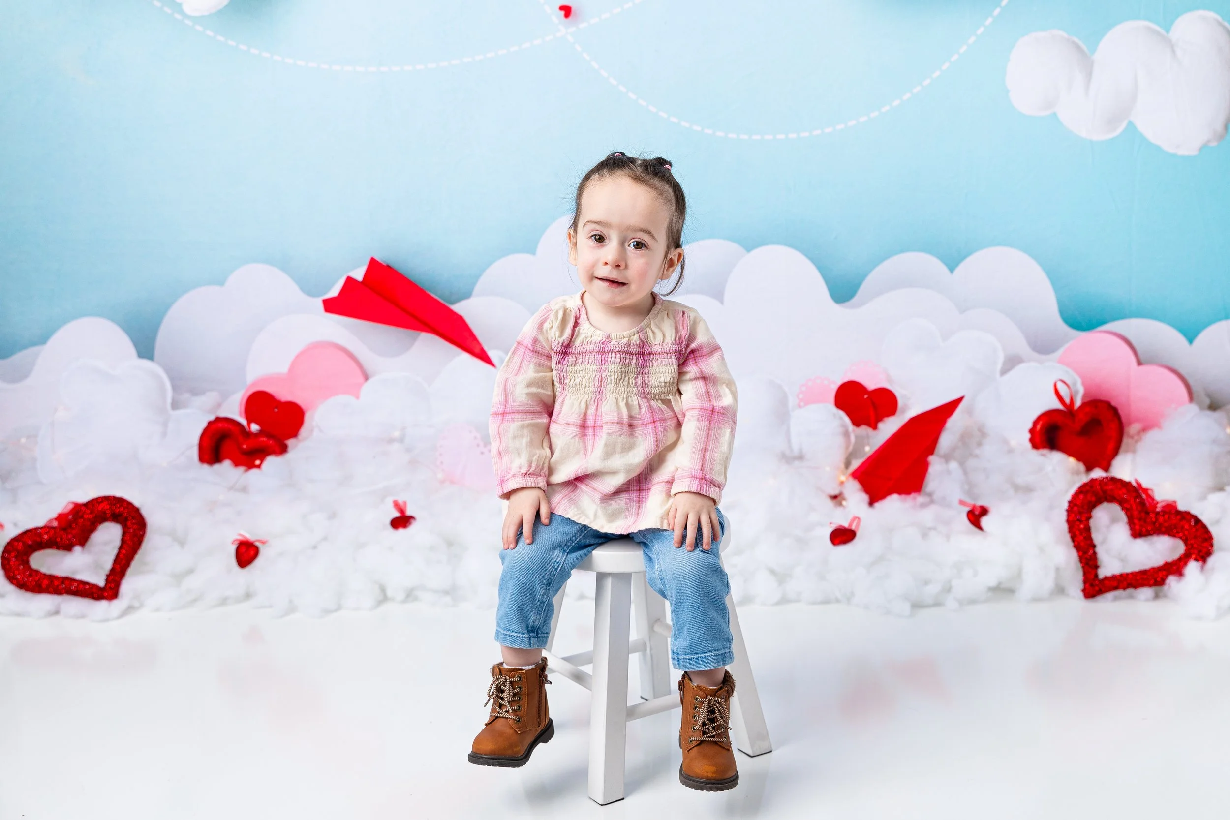 Young girl sitting on a white stool in front of a backdrop decorated with white clouds, red hearts, and red paper airplanes, with a blue sky background.