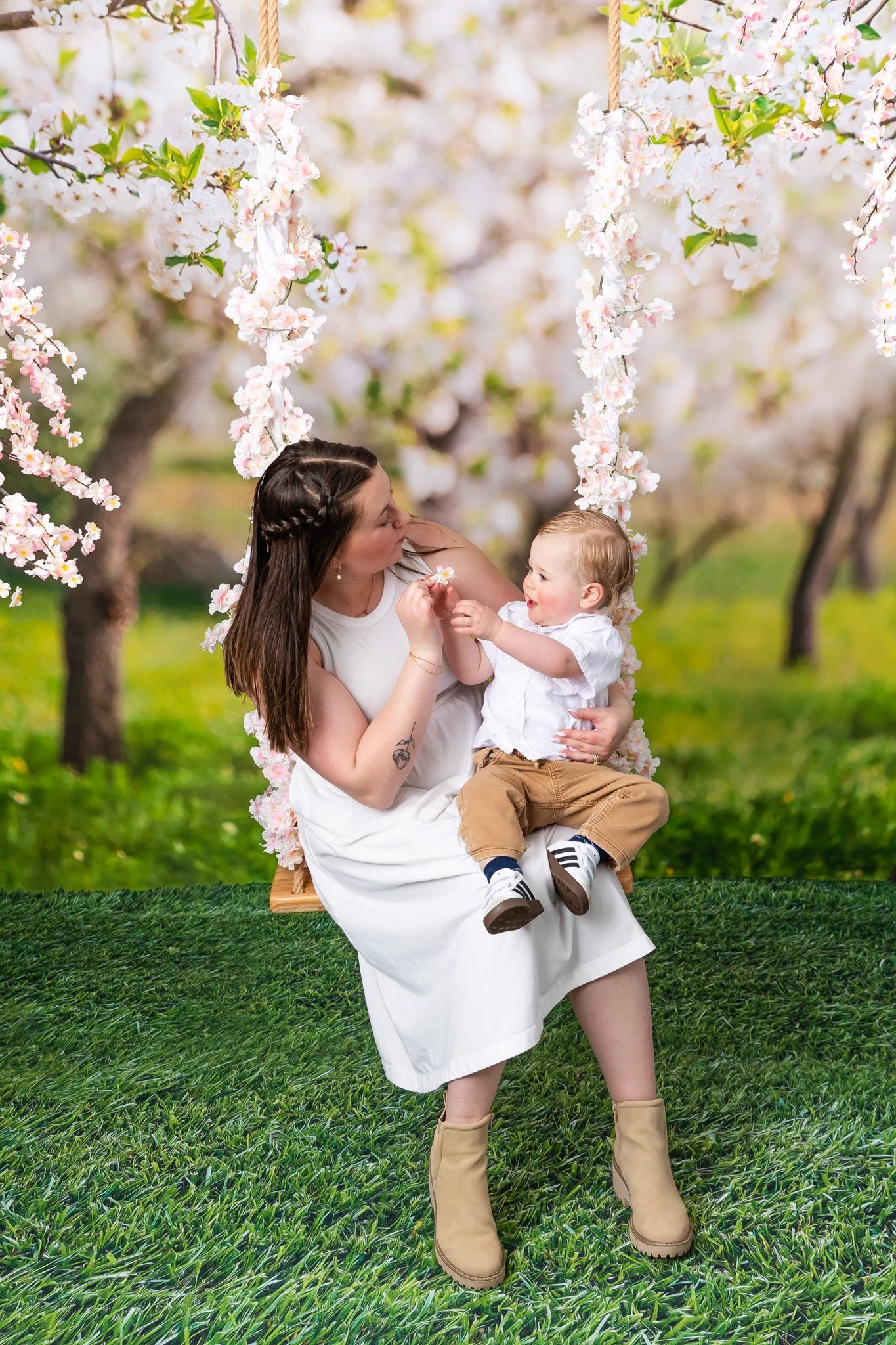 A woman and a young child sitting on a swing decorated with pink flowers, outdoors with blooming trees and green grass, sharing a moment of affection.
