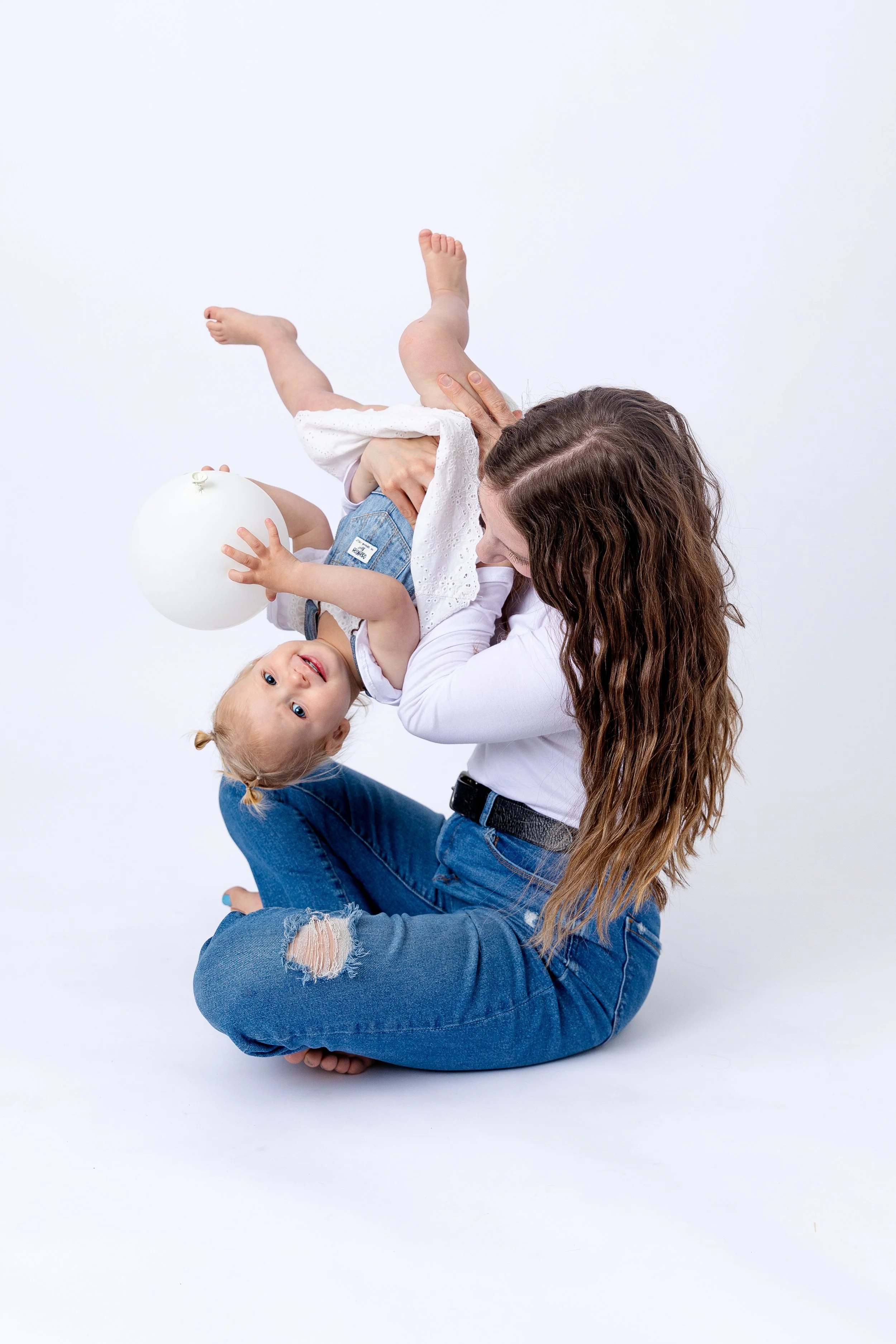 A woman with long curly brown hair sitting on the floor playing with a smiling blonde girl holding a white balloon, both dressed in casual white tops and blue jeans, with the girl upside down.