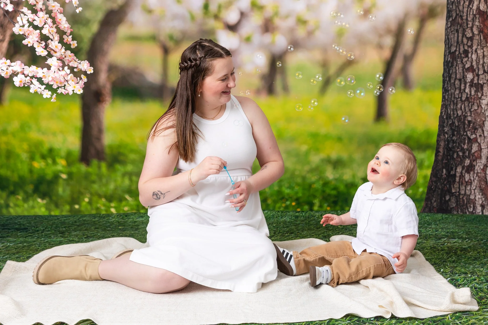 A woman and a young boy sitting on a blanket outdoors in a park with blooming trees, playing with bubbles and smiling at each other.