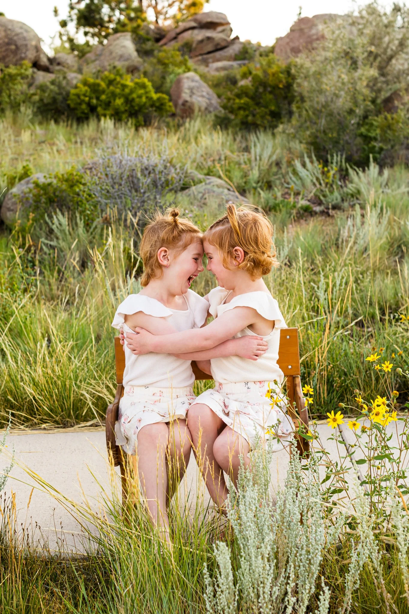 Two young children with red hair sitting on a wooden chair outdoors, smiling, and touching foreheads, surrounded by tall grass, yellow flowers, and bushes with a rocky hillside in the background.
