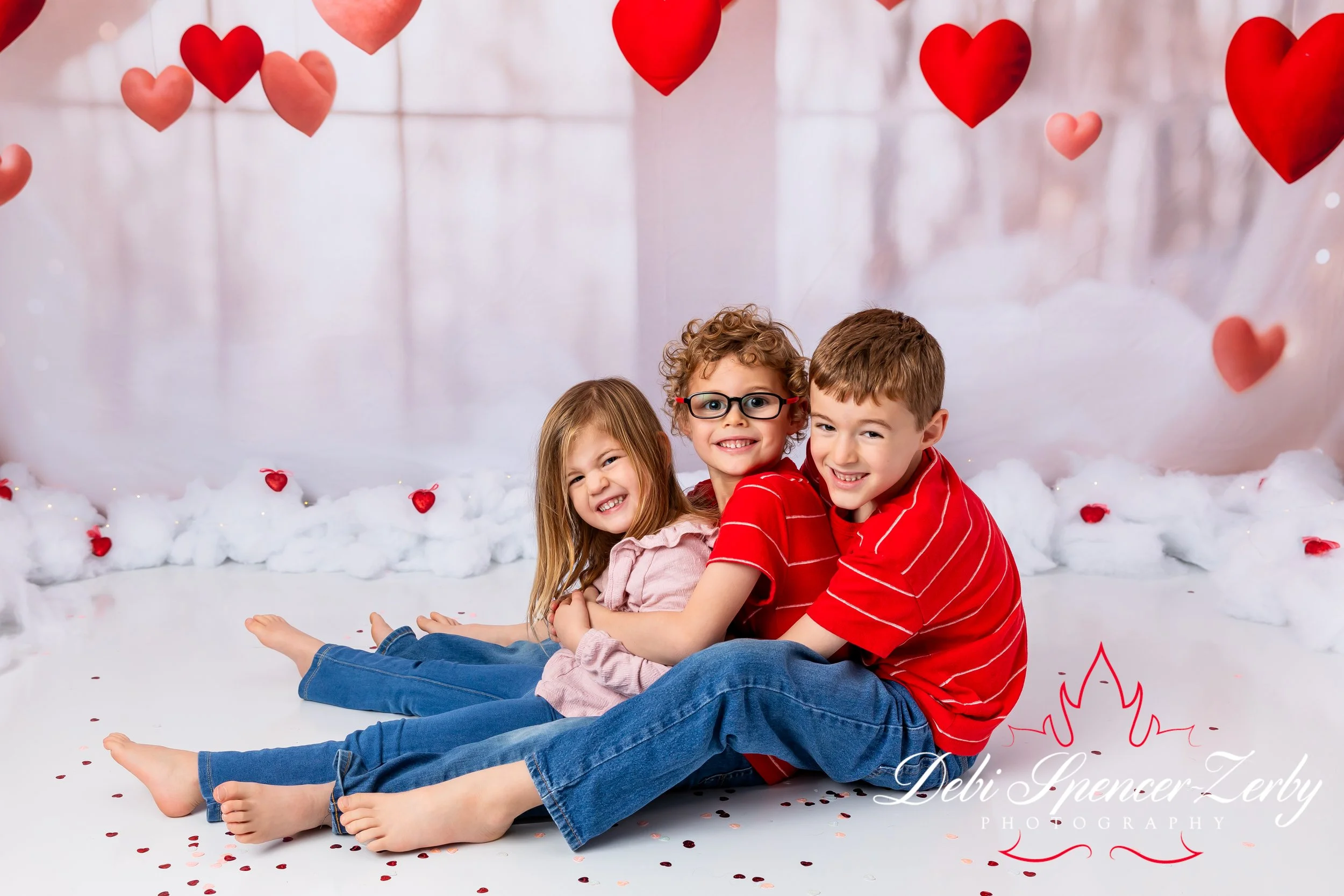 Three children sitting on the floor with attractions of red and pink hearts hanging from above and scattered on the fluffy white floor, with a backdrop of a light-colored wall.