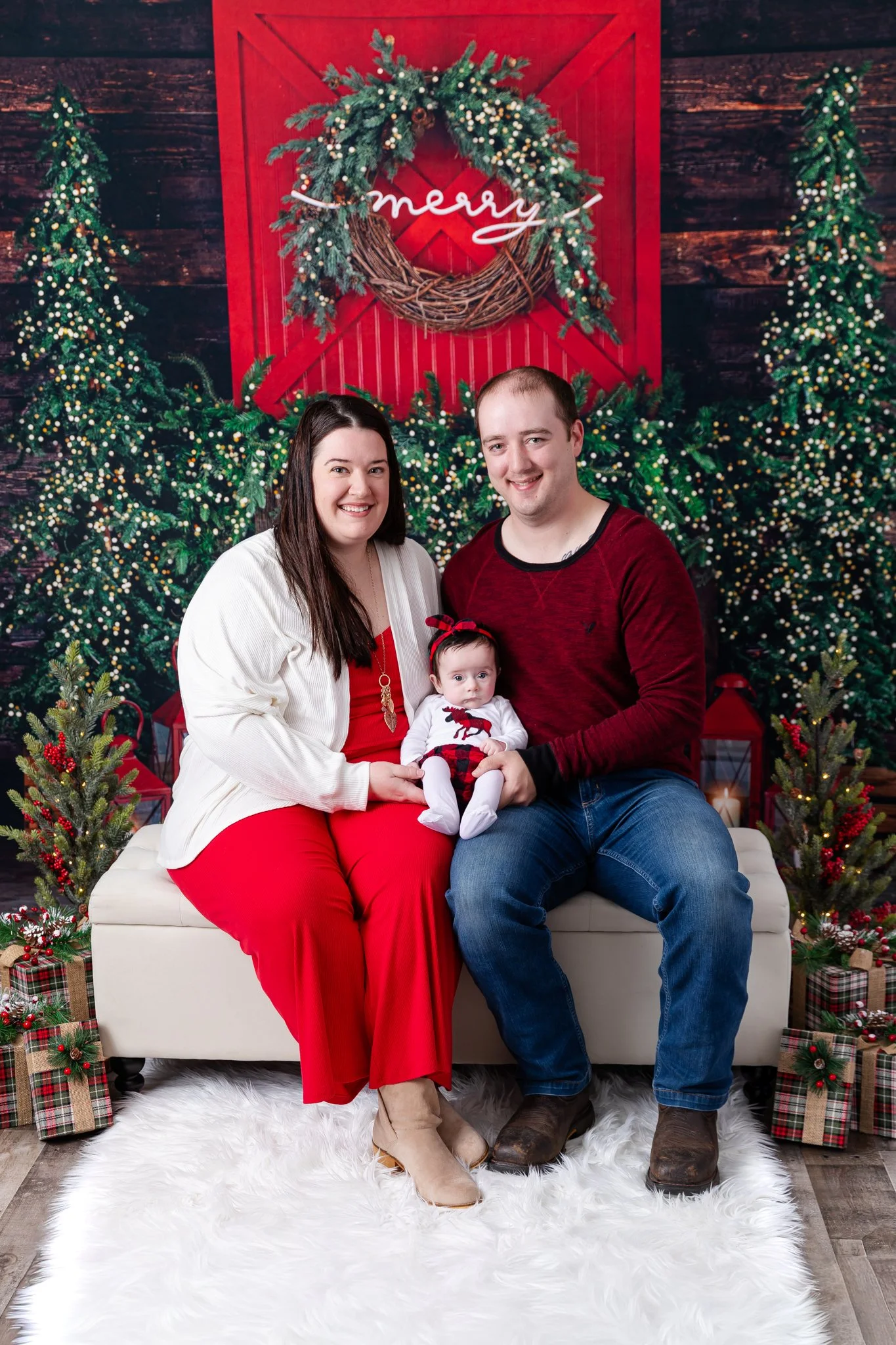 A family of three sitting on a white fluffy rug in front of a Christmas themed backdrop, including a large red barn door decorated with a wreath and the word 'merry' in neon lights, with Christmas trees, presents, and holiday decorations around them.