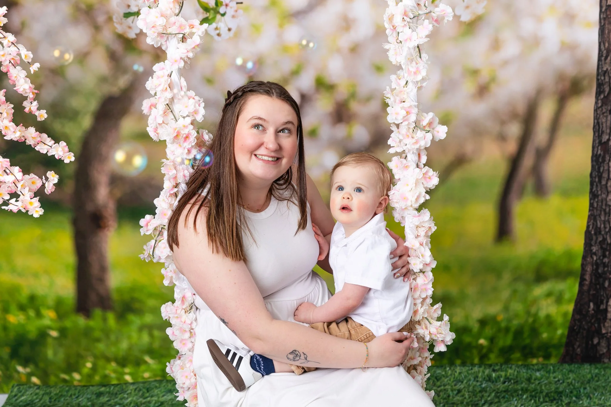 A woman and a young child sitting on a pink flower-decorated swing in a park with blossoming trees and green grass.