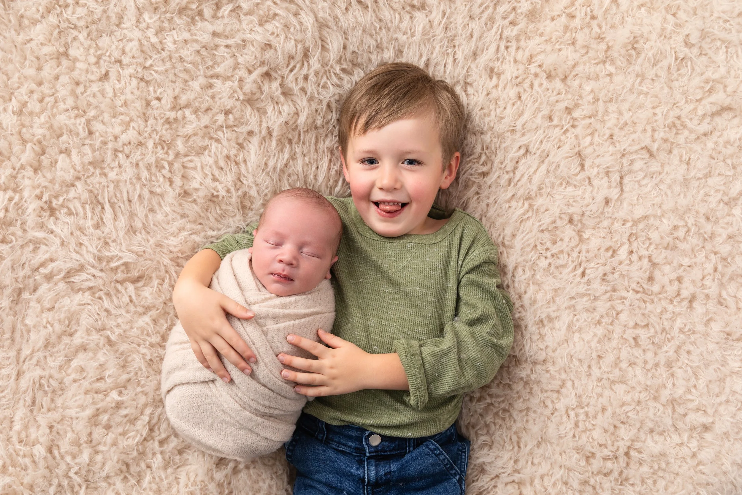 A young boy with brown hair and a green shirt holding a sleeping baby wrapped in a beige blanket on a soft, fluffy beige rug.