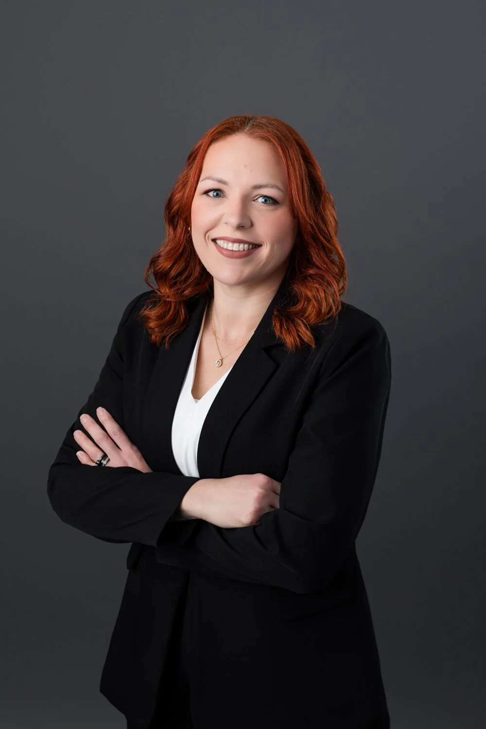 A professional woman with red hair and blue eyes smiling, wearing a black blazer and white shirt, with her arms crossed, against a dark gray background.