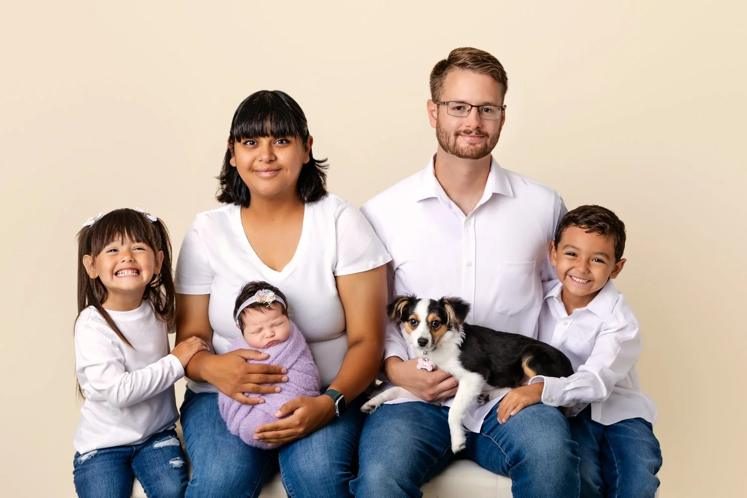 A family portrait featuring a woman holding a sleeping baby wrapped in a purple blanket, a girl with pigtails and a big smile, a man with glasses holding a dog, and a boy smiling, all sitting against a plain, light-colored background.