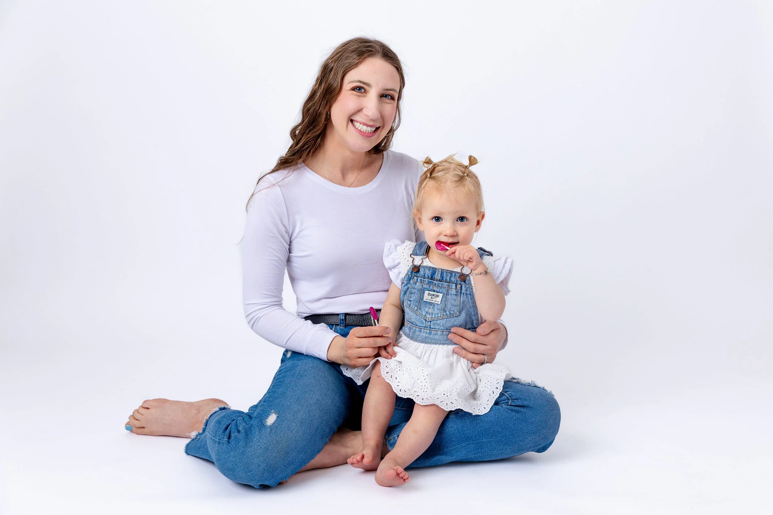 A smiling woman with long brown hair wearing a white long-sleeve shirt and ripped jeans, sitting cross-legged, holding a young girl with blonde hair in pigtails, wearing a denim dress over a white eyelet dress, and eating a lollipop.