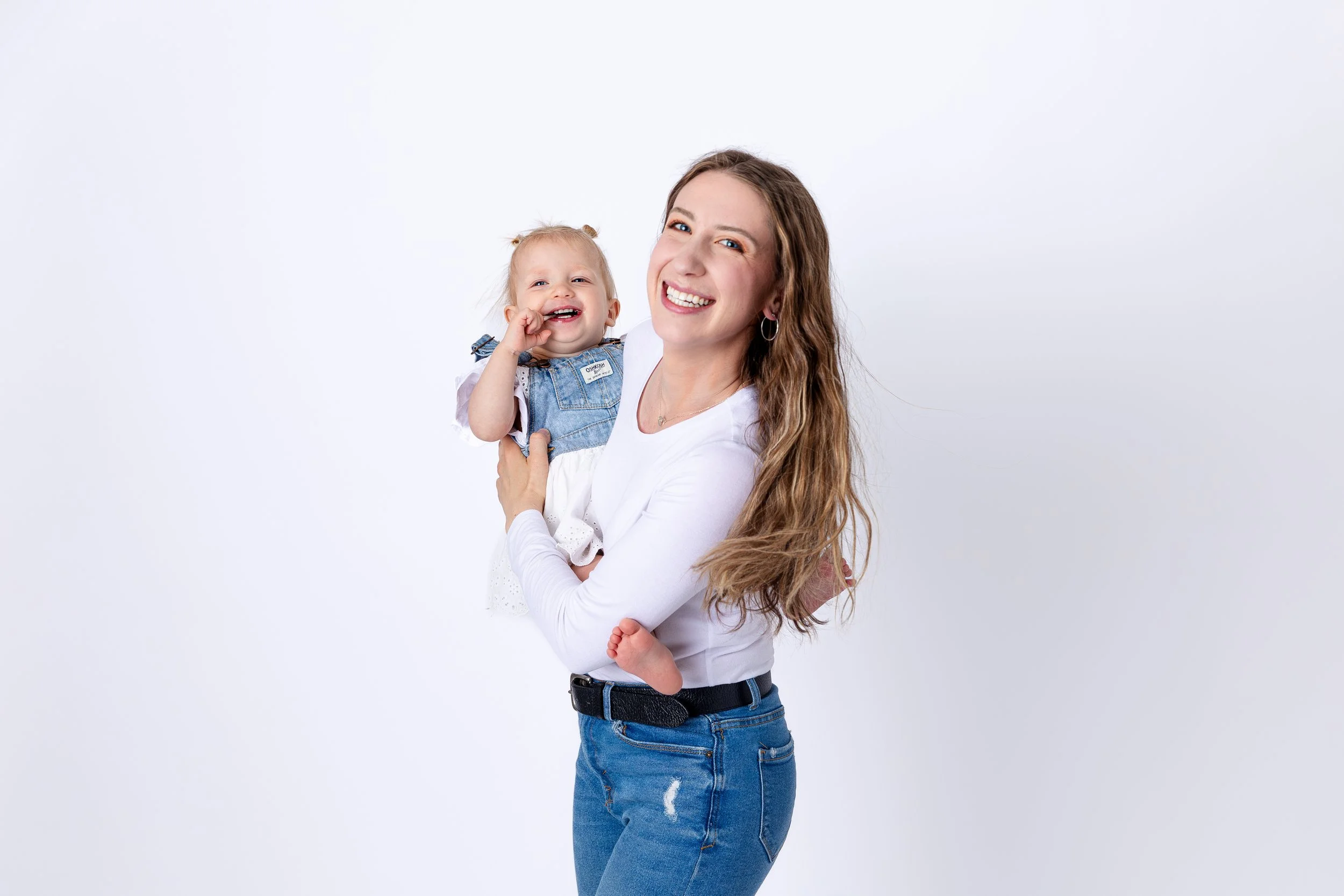 A woman with long wavy hair holding a smiling toddler girl with blonde hair in pigtails, both dressed casually against a plain white background.