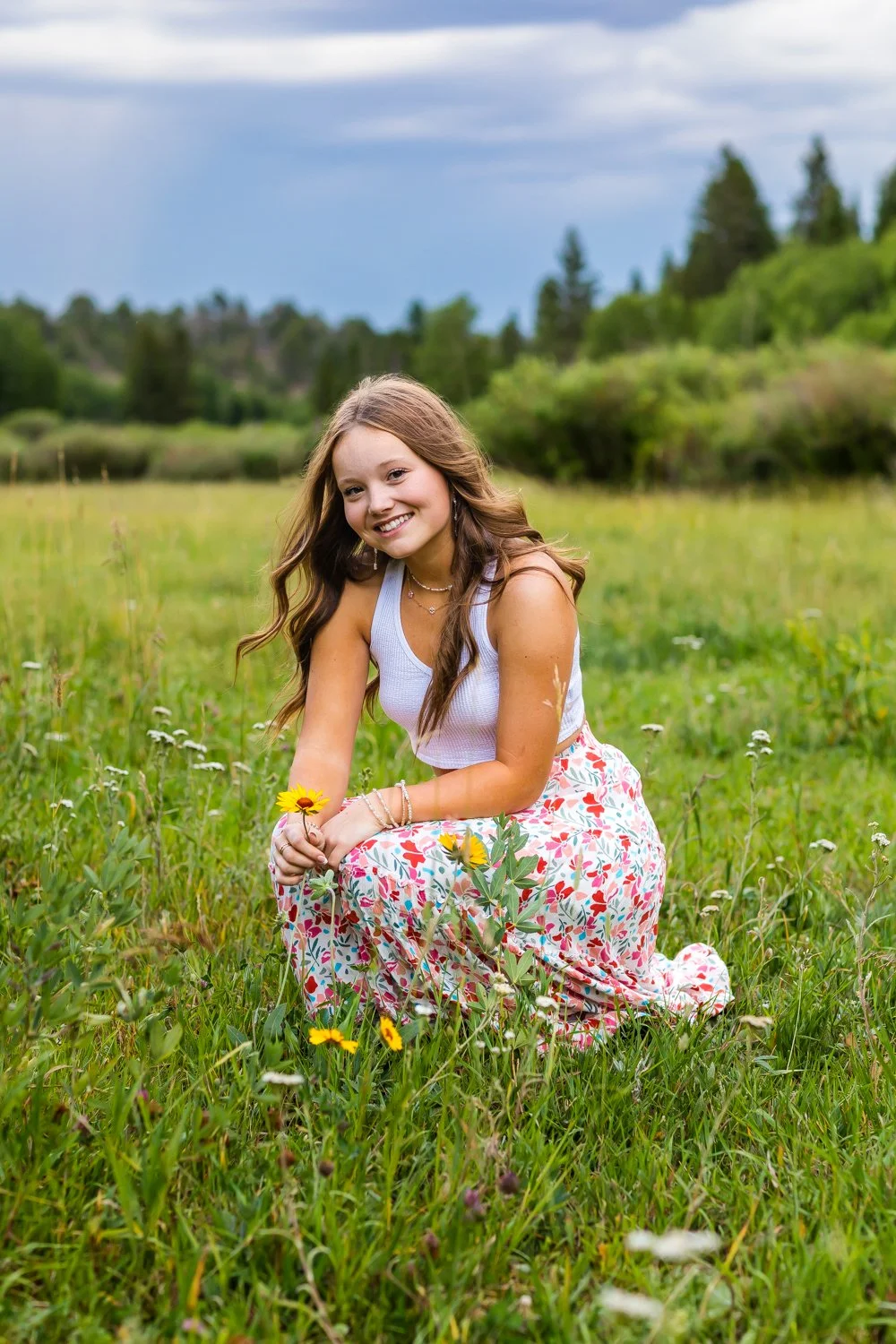 Young woman with long wavy hair smiling while kneeling in a grassy field holding a yellow flower.