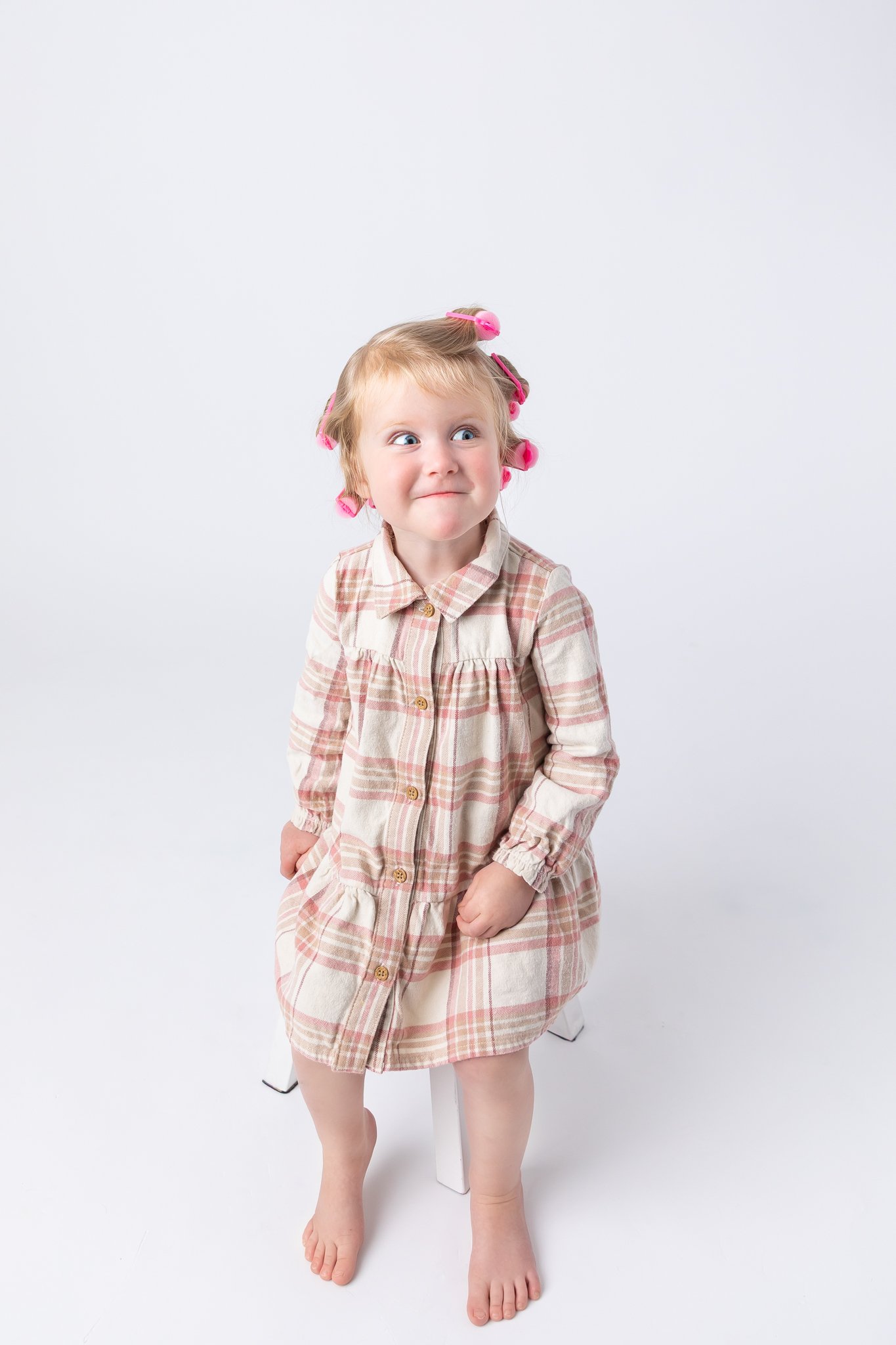 A young girl with pink hair rollers in her hair, sitting on a white stool, making a funny face, wearing a beige and pink plaid dress.