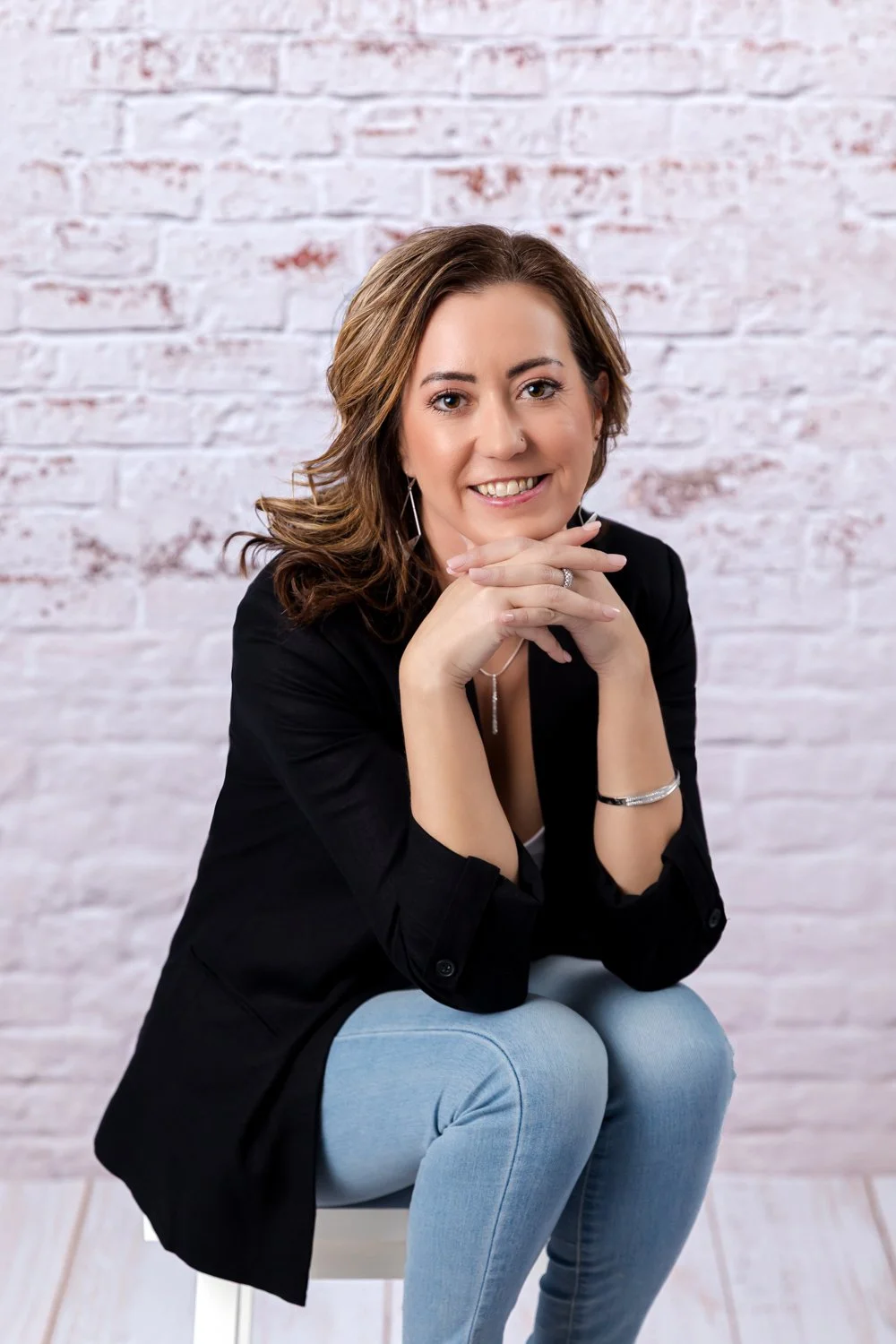 A woman with brown hair smiling, sitting with her chin resting on her hands, wearing a black blazer and light blue jeans, posed in front of a white brick wall.