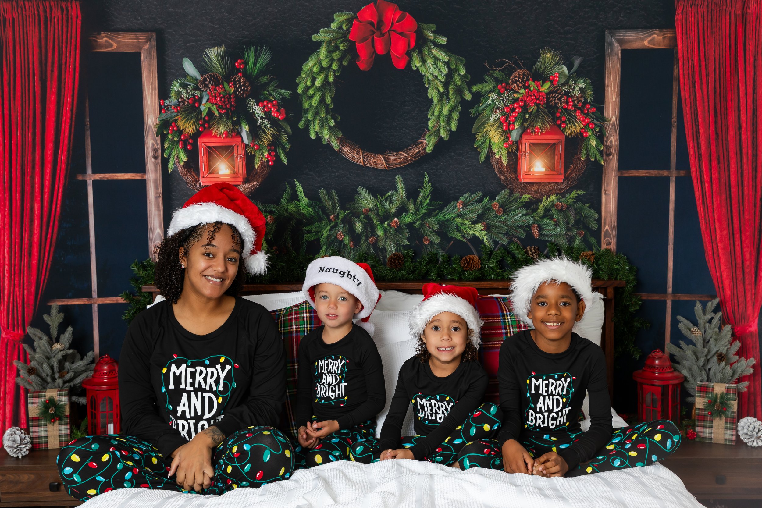 A woman and four children sitting together on a bed, wearing black pajamas with holiday messages and Christmas hats, in front of a festive holiday backdrop with wreaths, red curtains, and Christmas decorations.