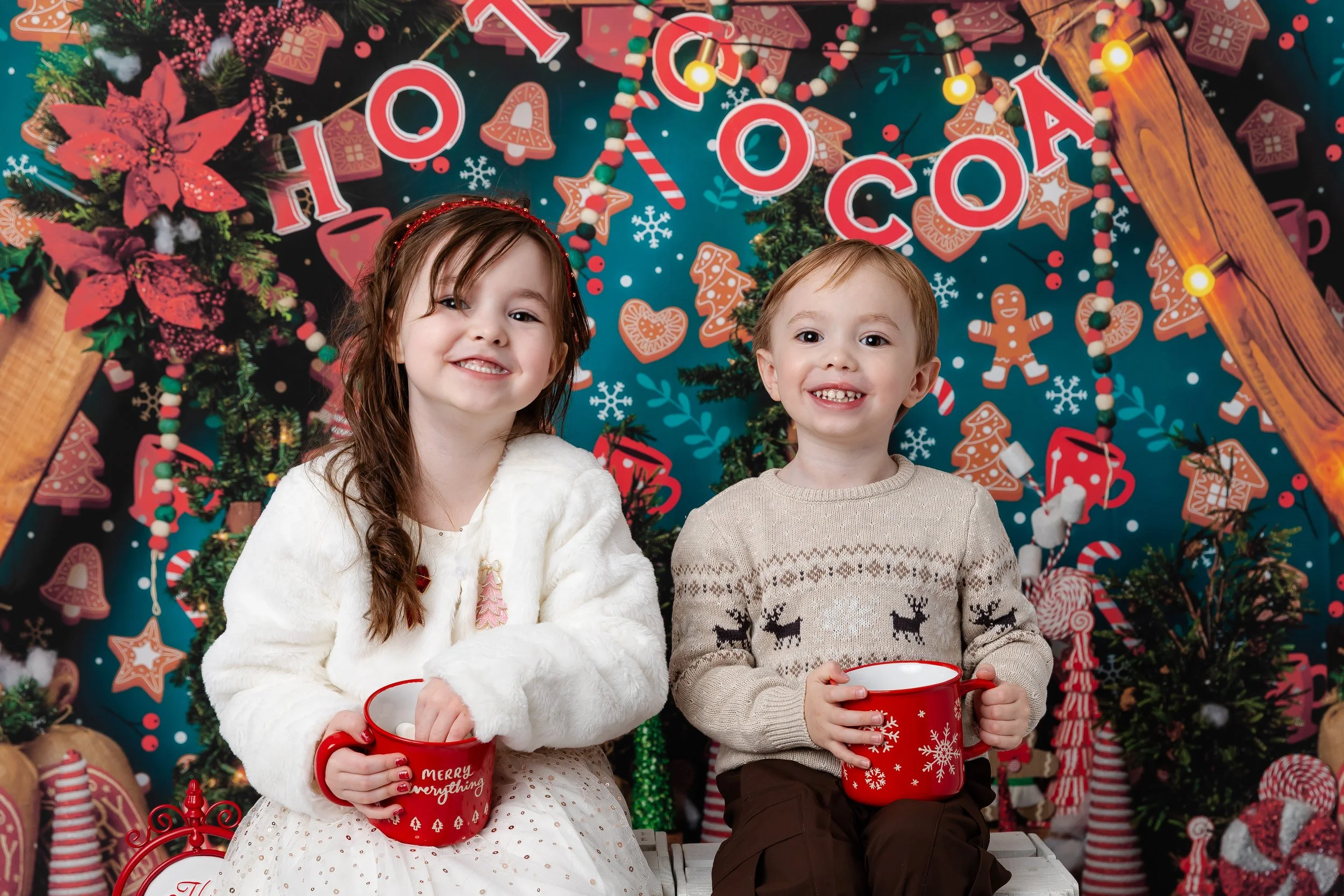 Two children sitting with Christmas-themed red mugs, smiling, in front of a festive holiday backdrop featuring Christmas decorations, gingerbread cookies, and fairy lights.