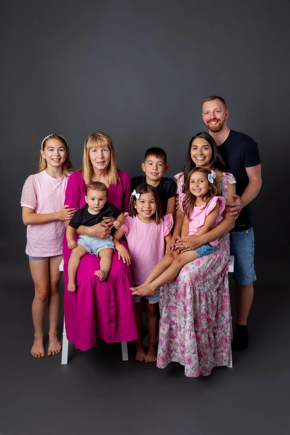 A multigenerational family portrait with seven members, including children, a woman, and two men, smiling and posing against a dark gray background, some sitting on stools or standing.