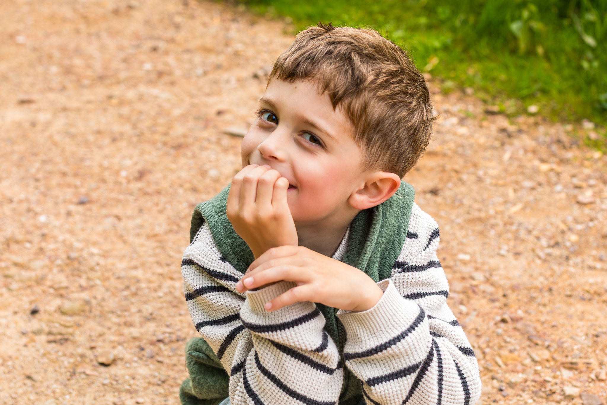 A young boy with brown hair and blue eyes smiling while sitting outdoors on a dirt path with green grass to the right.