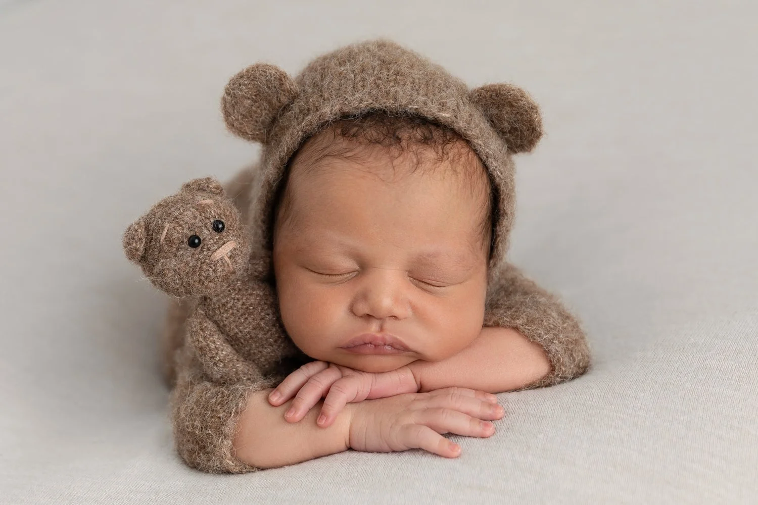 A sleeping baby dressed in a brown bear costume, including a hat with bear ears, resting on a white surface. The baby has closed eyes and appears peaceful.