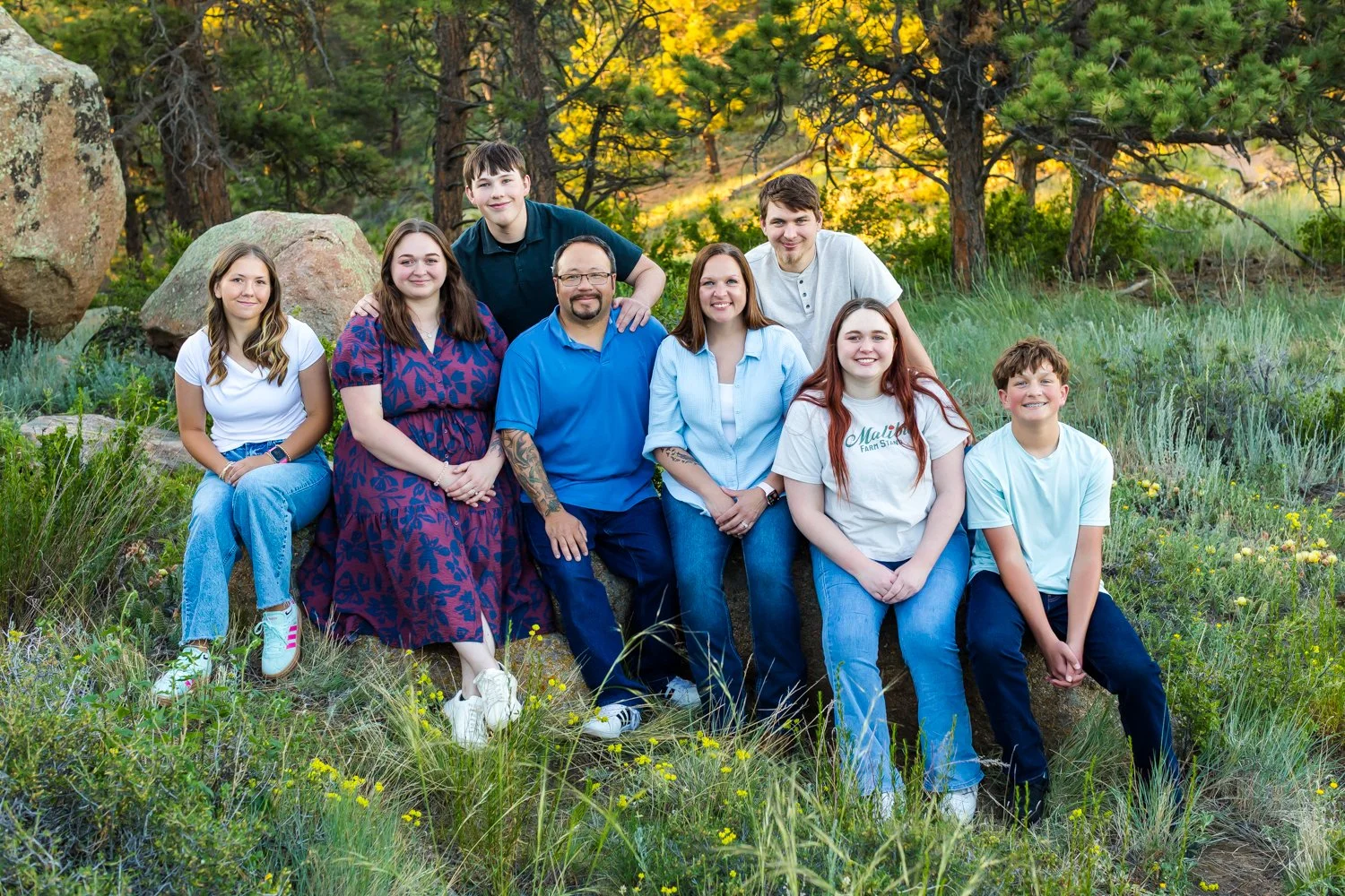 A group of nine people posing outdoors in a grassy area with trees and rocks in the background. They are smiling and sitting or standing close together.