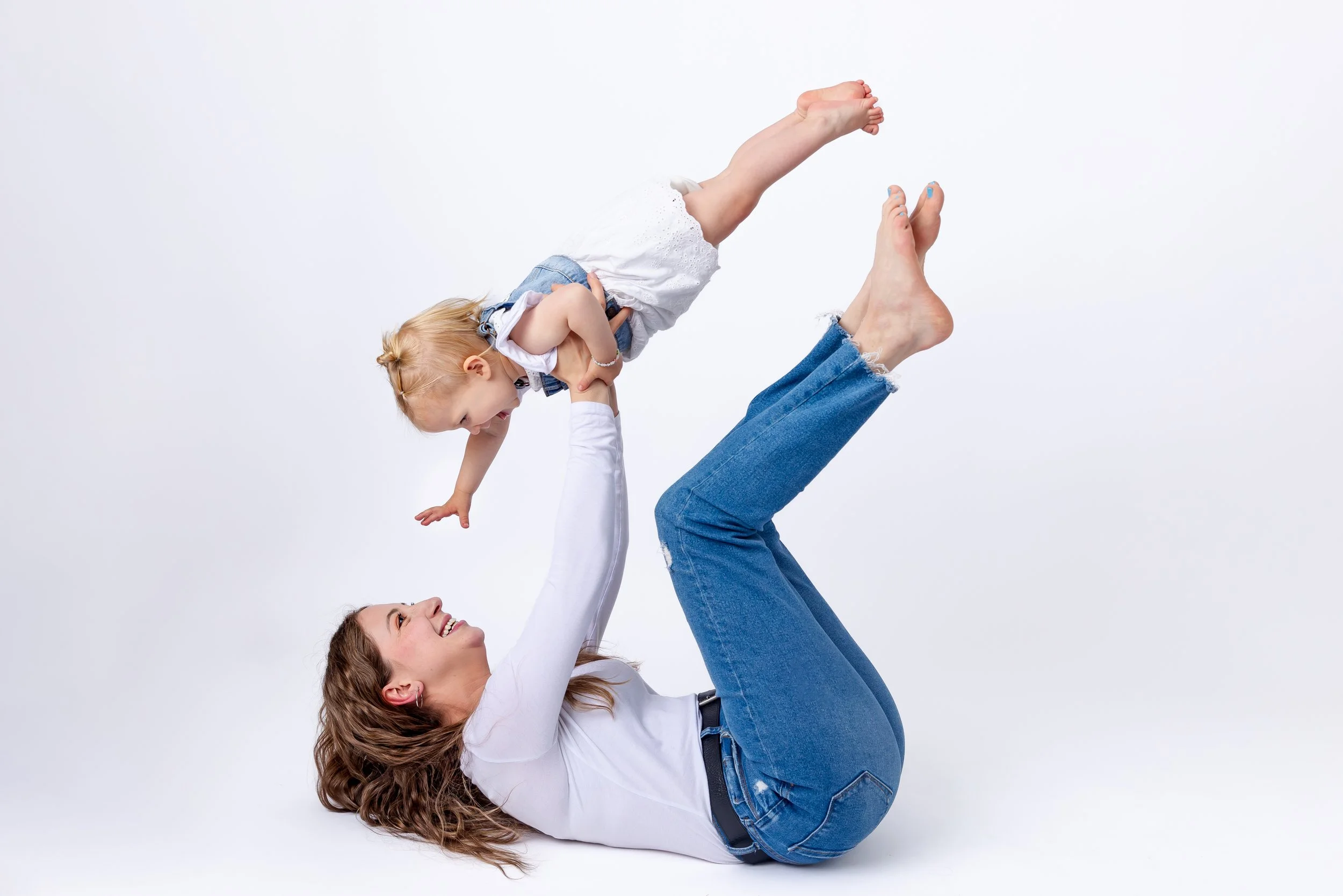 A woman lying on her back lifting a young girl in the air, both smiling, against a plain white background.